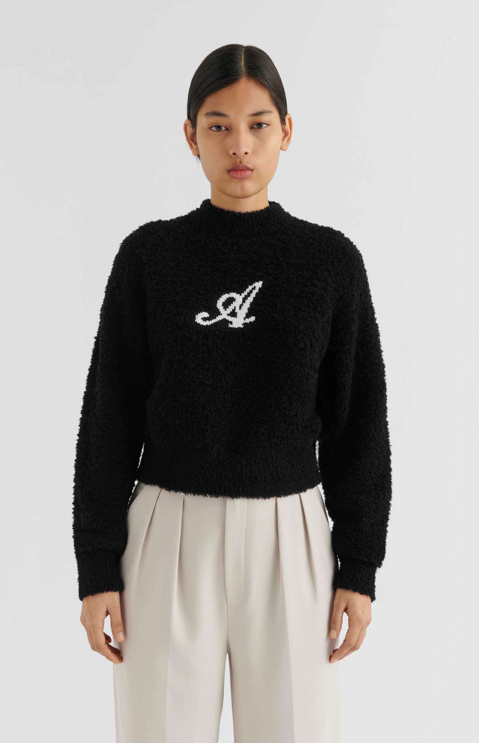 A young woman with dark hair stands against a white background, wearing a black, fuzzy, high-neck sweater with a white cursive 'A' logo on the chest, and light beige pleated trousers. She looks directly at the viewer with a neutral expression.