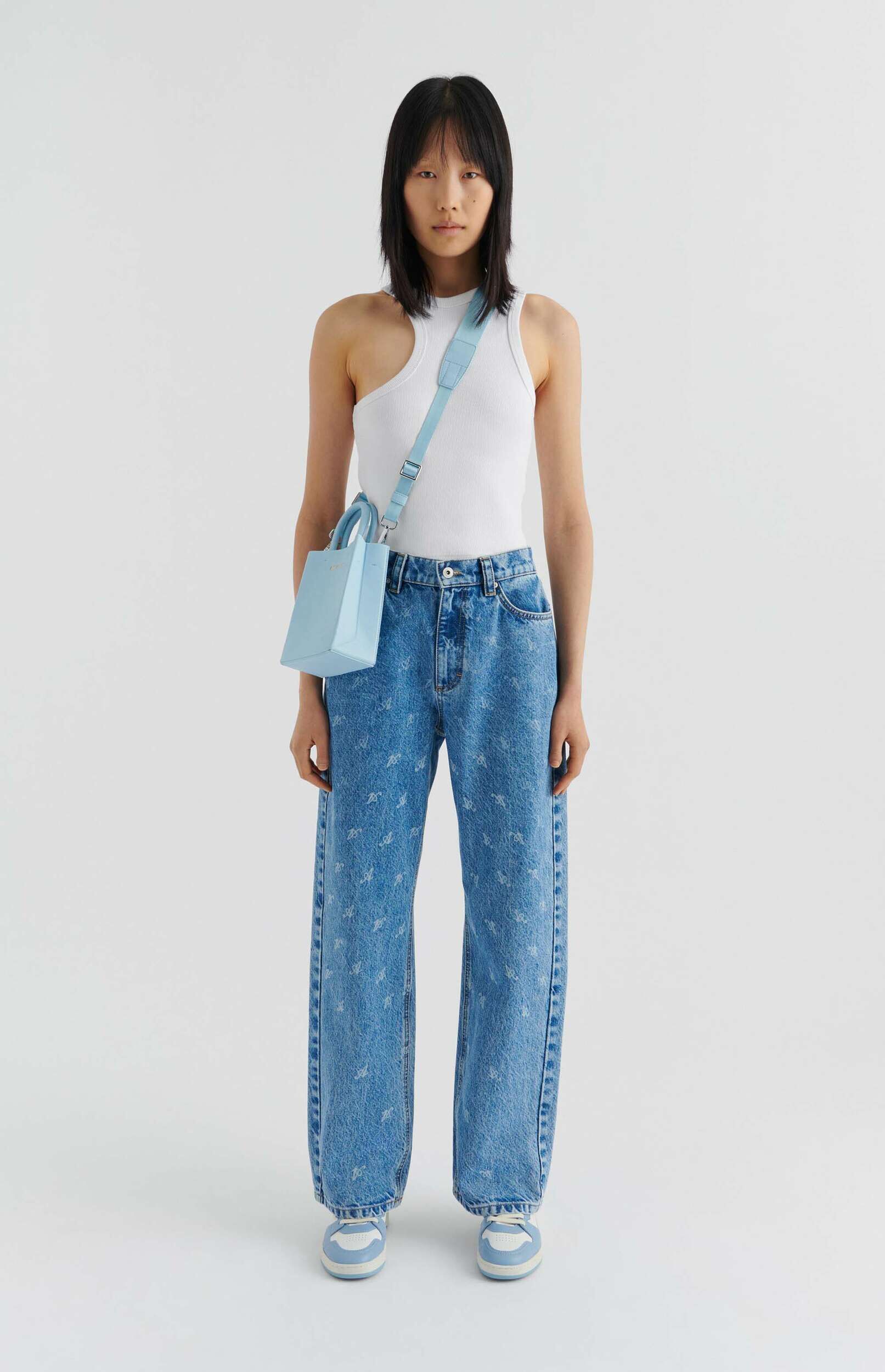 A full-body shot of a person standing against a white background, wearing a white ribbed tank top, light blue wide-leg jeans with a subtle light-colored pattern, and light blue and white sneakers. A small light blue shoulder bag is worn across their body. The person has dark, chin-length hair and is looking directly at the camera.