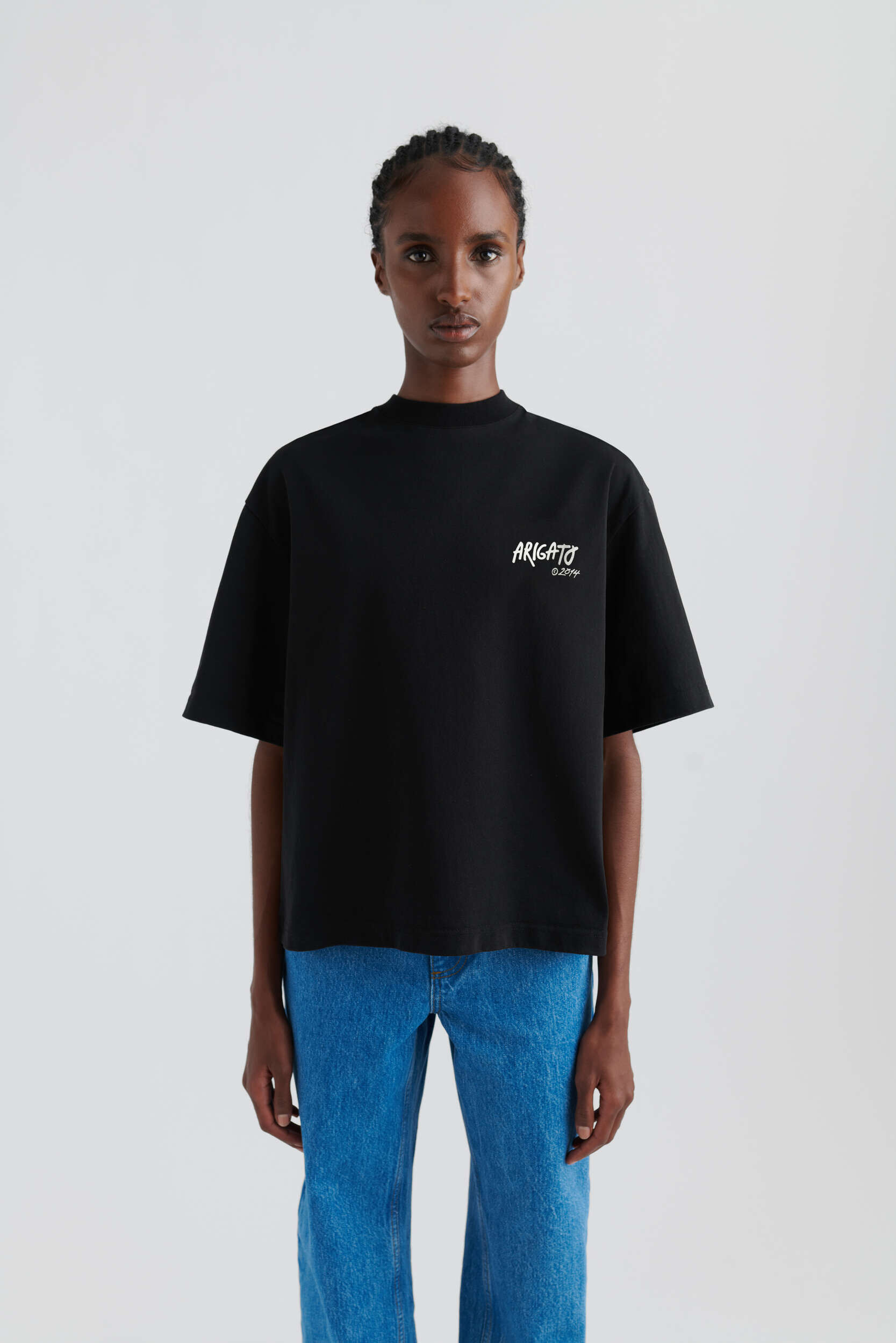 A dark-skinned model with cornrow braids wears a black short-sleeved t-shirt with 'ARIGATO ©2014' printed in white on the chest, paired with blue jeans, standing against a light background.