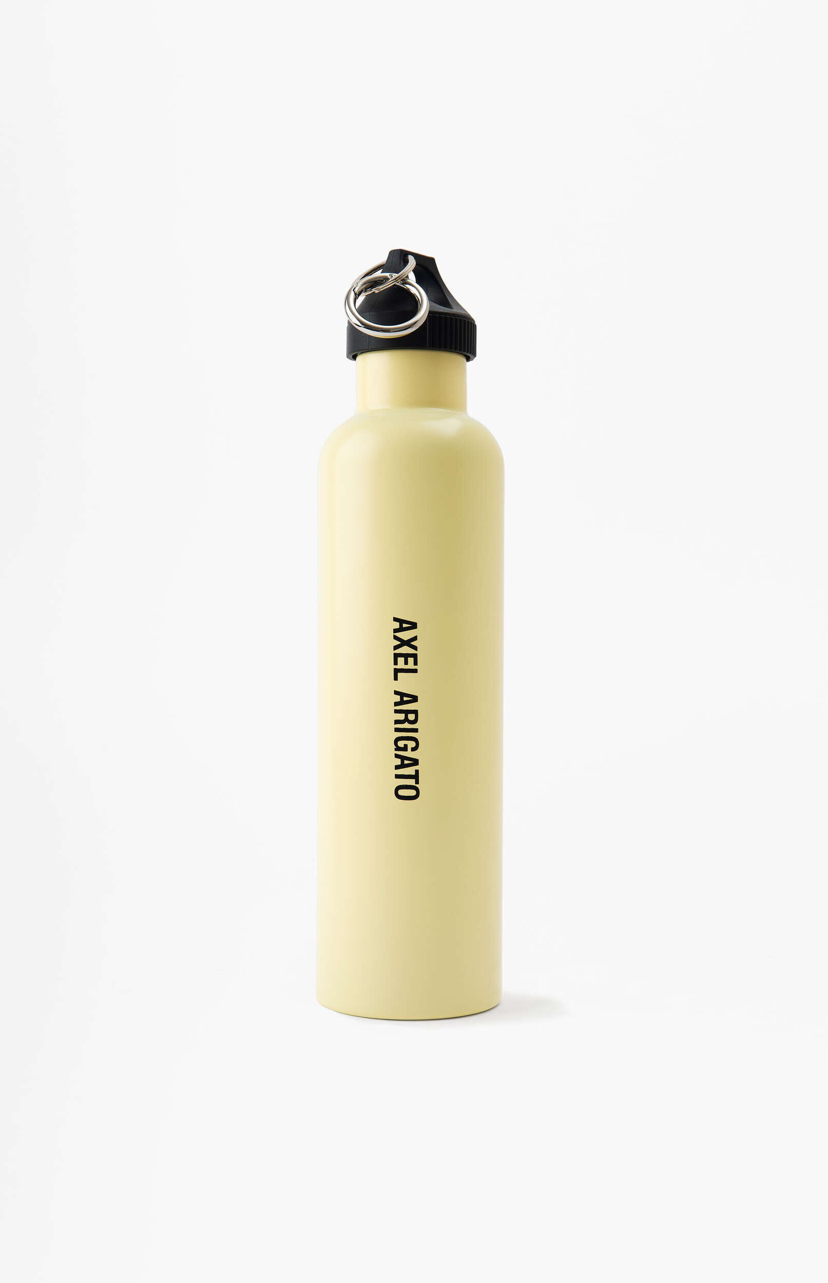 A light yellow reusable water bottle by Axel Arigato stands upright against a clean white background. The bottle has a matte finish, a black screw-on cap, and a metal split ring attached to the cap. The brand name 'AXEL ARIGATO' is printed vertically in black letters on the side of the bottle.