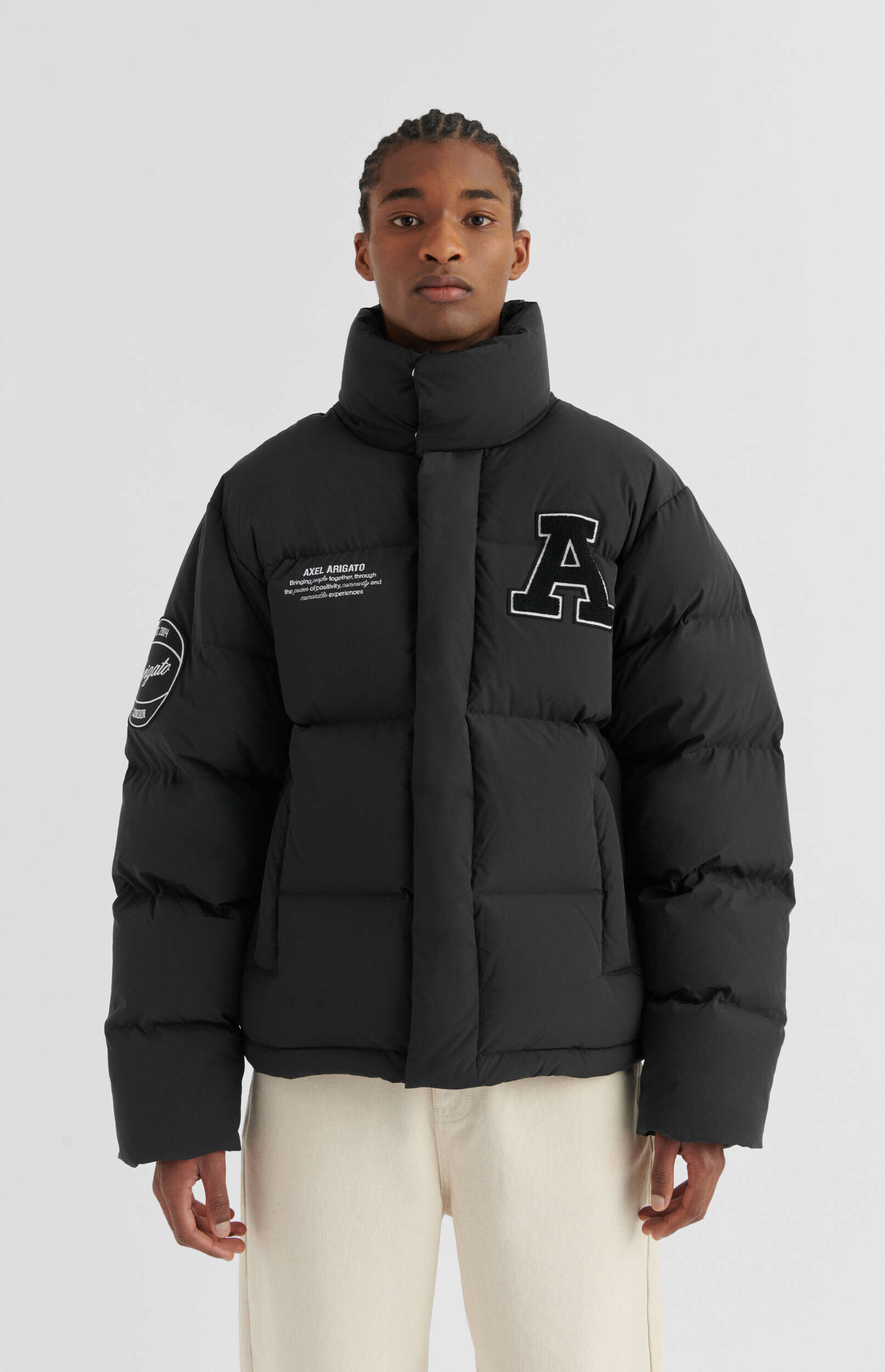 A man with braided hair stands facing forward, wearing a black puffer jacket with a high collar and light cream pants. The jacket features a large white letter 'A' with a black outline on the right chest, and 'AXEL ARIGATO' text on the left chest. A circular patch is visible on the left sleeve.