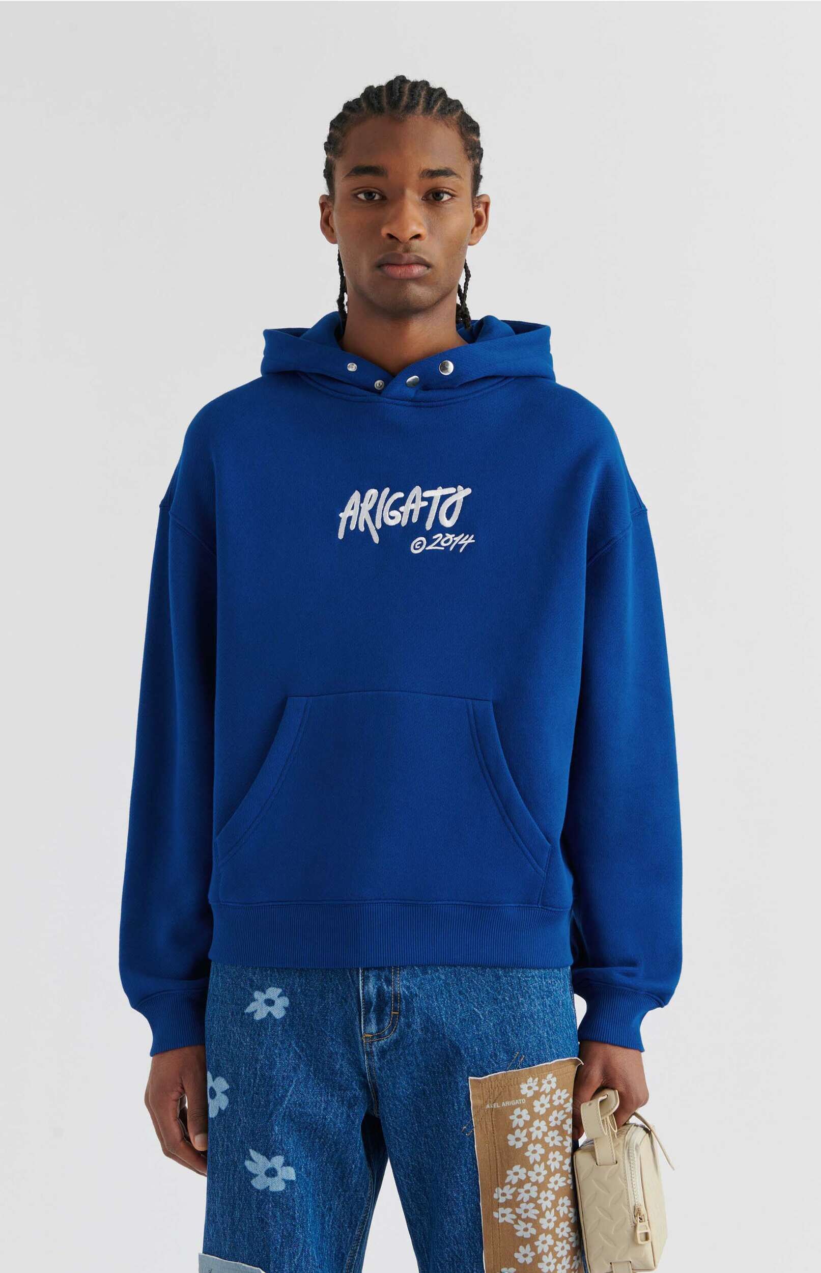 A person wearing a vibrant royal blue hoodie with "ARIGATO ©2014" written in white, blue jeans featuring white floral patterns, and holding a small patterned beige bag along with a brown fabric swatch printed with white flowers and "AXEL ARIGATO" text. The person has braided hair and is looking directly forward against a white background.