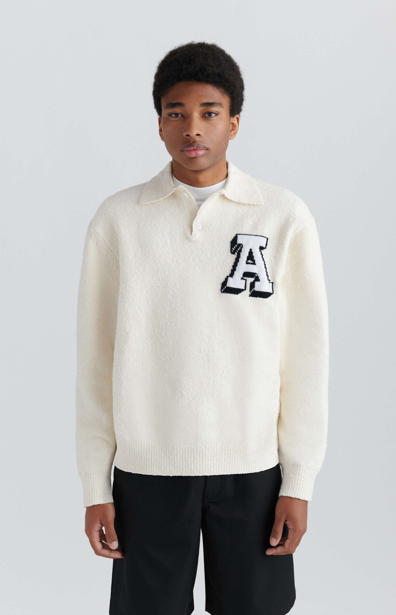 A young man with dark curly hair and a neutral expression stands against a plain white background, wearing a cream-colored long-sleeve polo sweater with a large black and white letter 'A' patch on the left chest, and black shorts.
