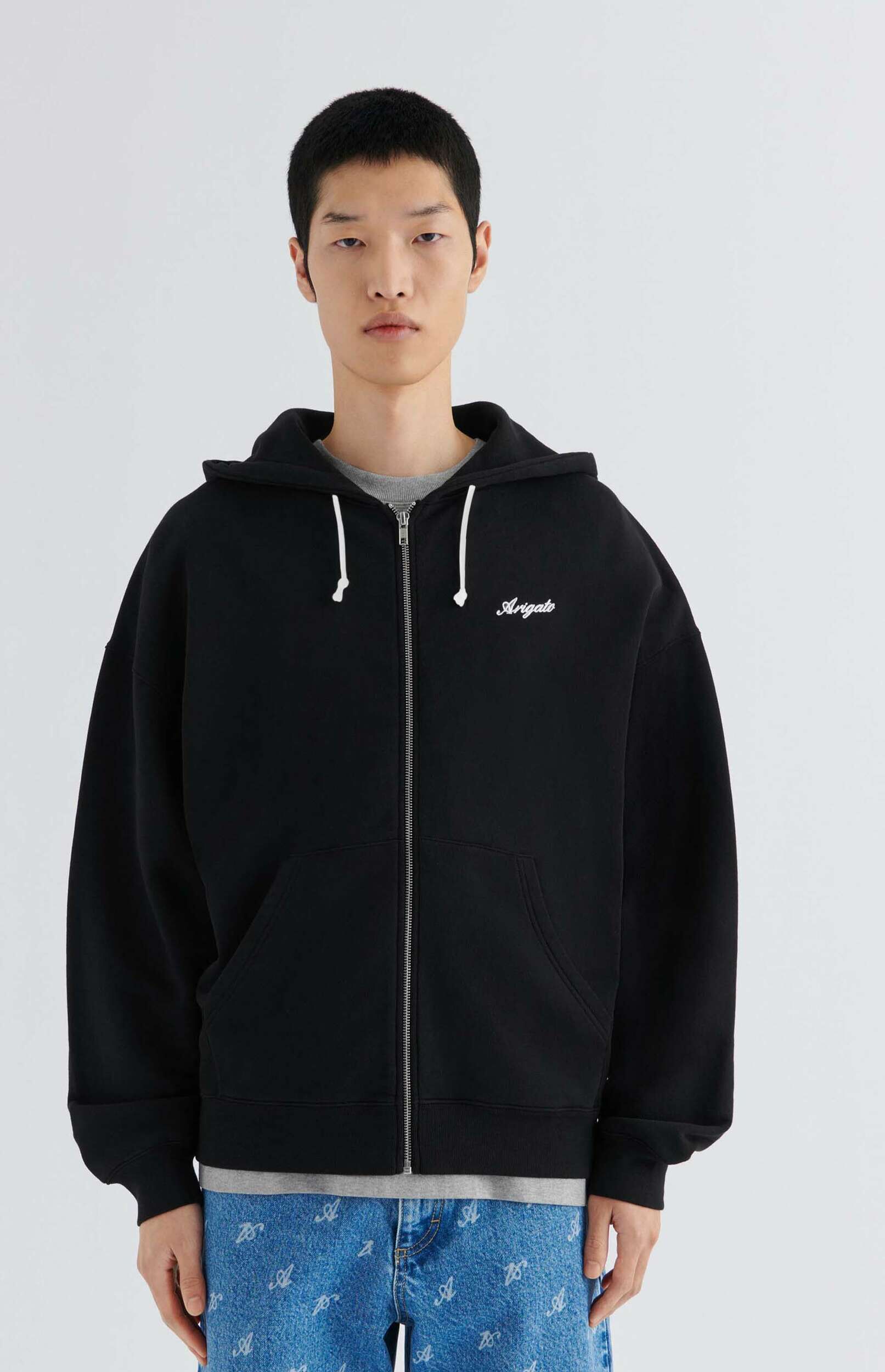 A young man models a black zip-up hoodie featuring 'Arigato' in white script on the chest, paired with a grey t-shirt and light blue jeans adorned with a white repeating 'A' pattern. He stands against a clean white background.