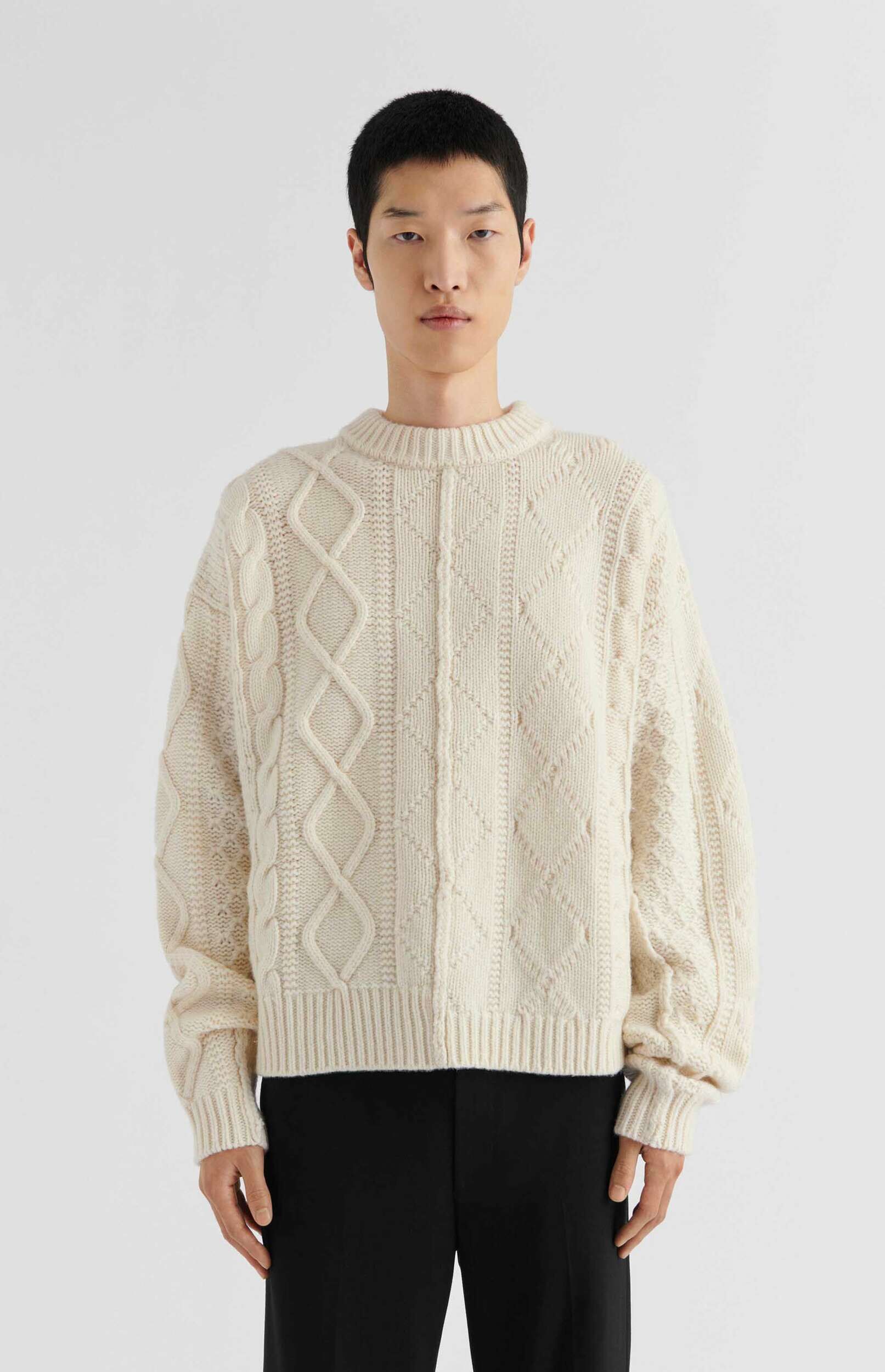 A male model with short black hair is wearing a cream-colored cable knit sweater and black trousers, standing against a plain white background. The sweater features various knit patterns, including prominent cable braids, diamond patterns, and ribbed details on the collar, cuffs, and hem. The model's hands are relaxed by his sides, and he looks directly at the viewer.
