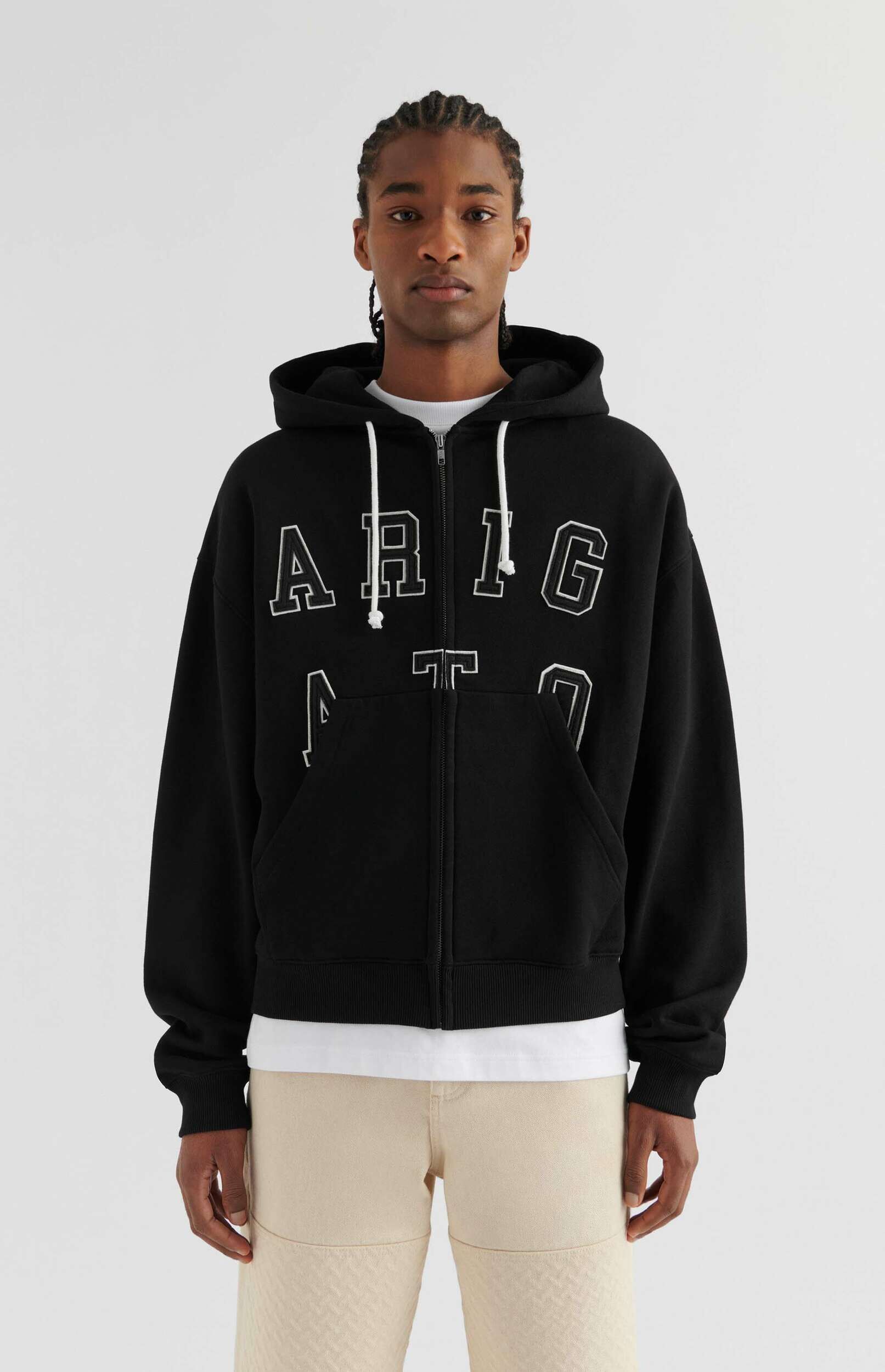 A young man with braided hair stands facing forward, wearing a black zip-up hoodie with white outlined letters 'ARIG' and a white t-shirt underneath, paired with light beige pants against a clean white background.
