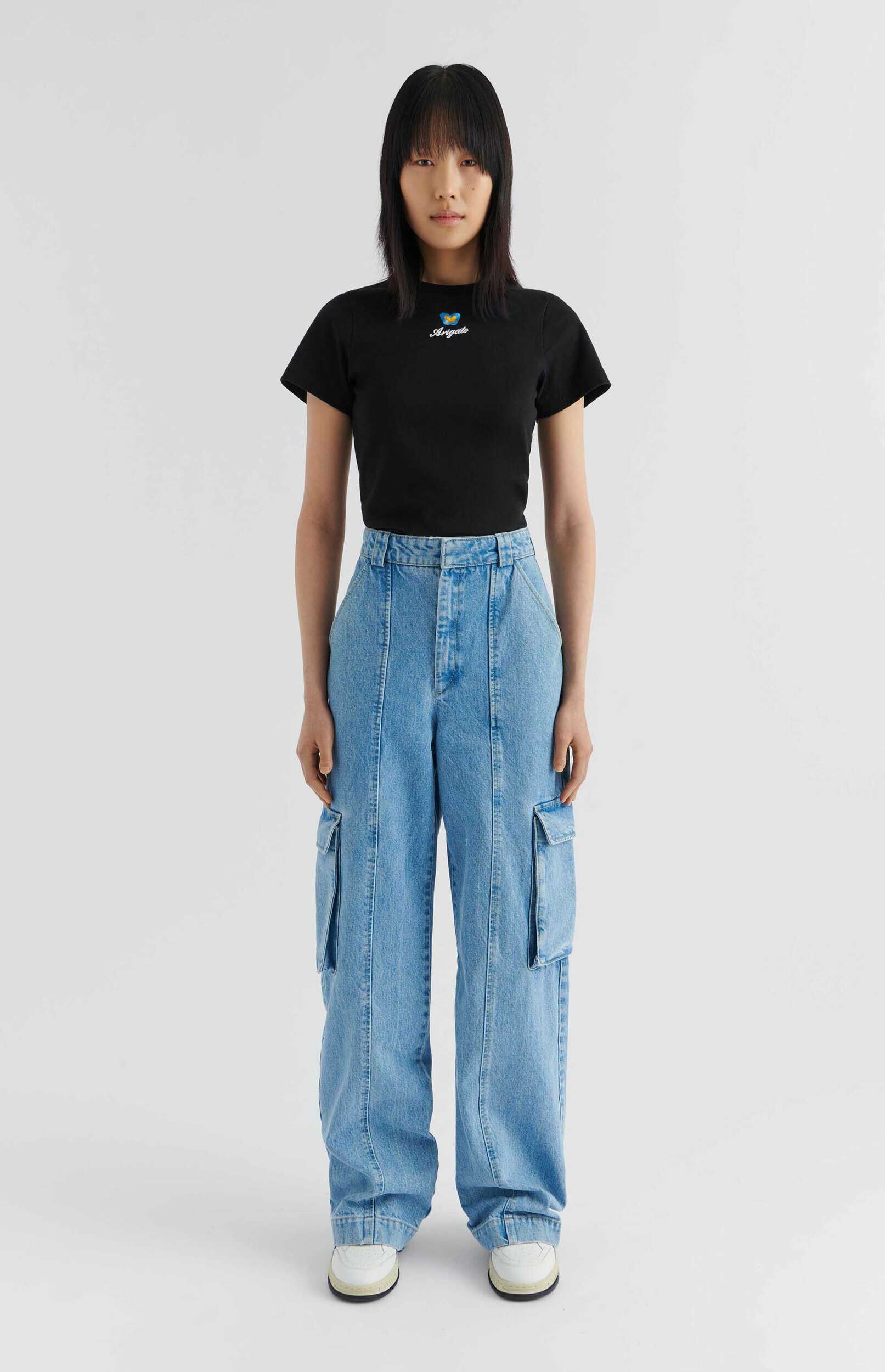 A full-body shot of a person standing against a white background, wearing a black t-shirt with a blue and yellow butterfly design and 'Arigato' text, paired with light blue wide-leg denim cargo pants and white sneakers.