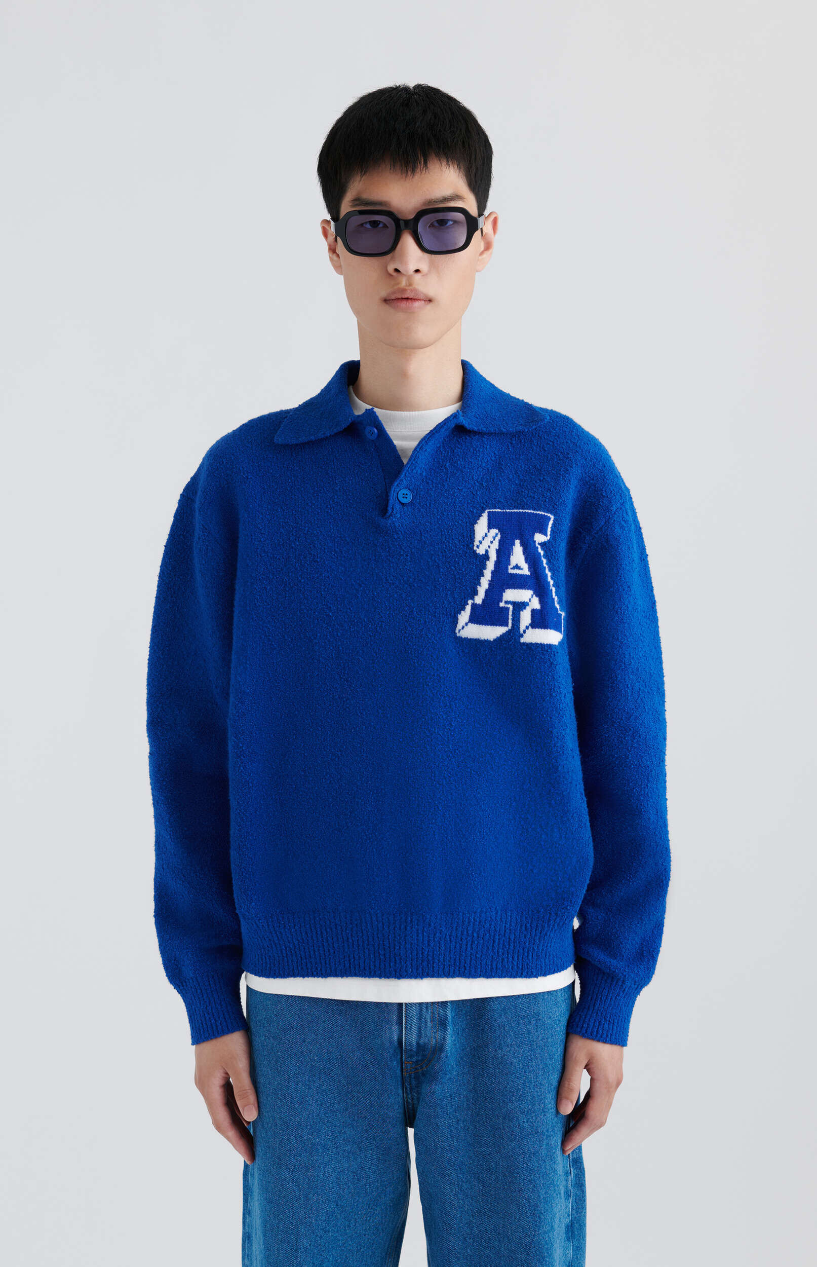 A man wearing a royal blue knitted polo sweater with a white outlined 'A' logo on the chest, light blue jeans, and rectangular black sunglasses, standing against a plain white background.