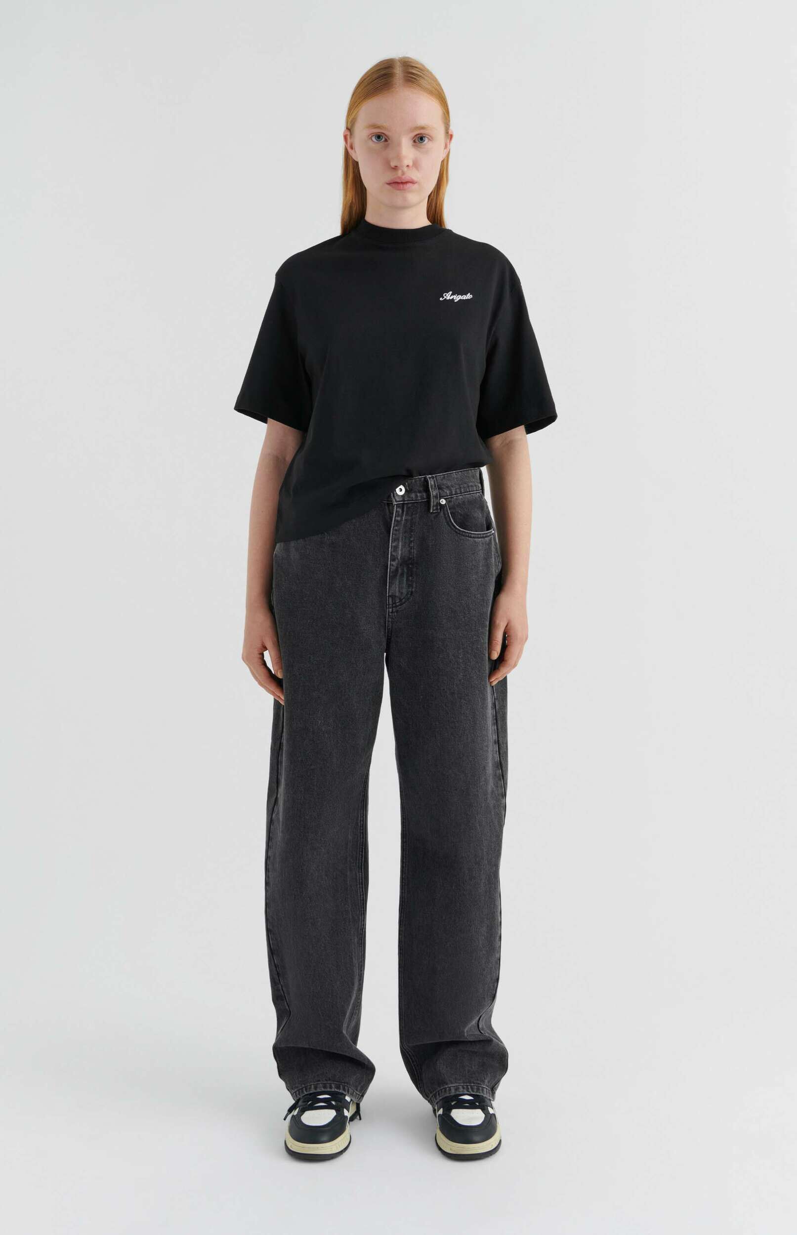 A full-body shot of a young woman with light brown hair wearing a black t-shirt with 'Arigato' in white script, dark gray relaxed-fit jeans, and black and white sneakers, standing against a white background.