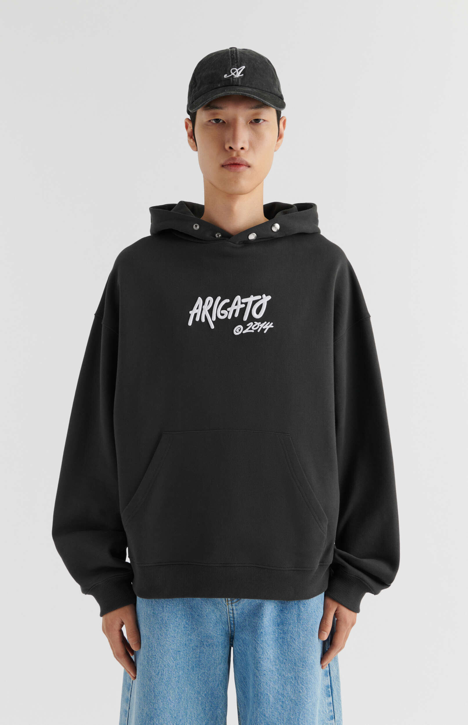 A male model stands against a plain light background, wearing a black oversized hoodie with 'ARIGATO © 2014' printed in white script, light blue wide-leg jeans, and a distressed black baseball cap with a white logo.