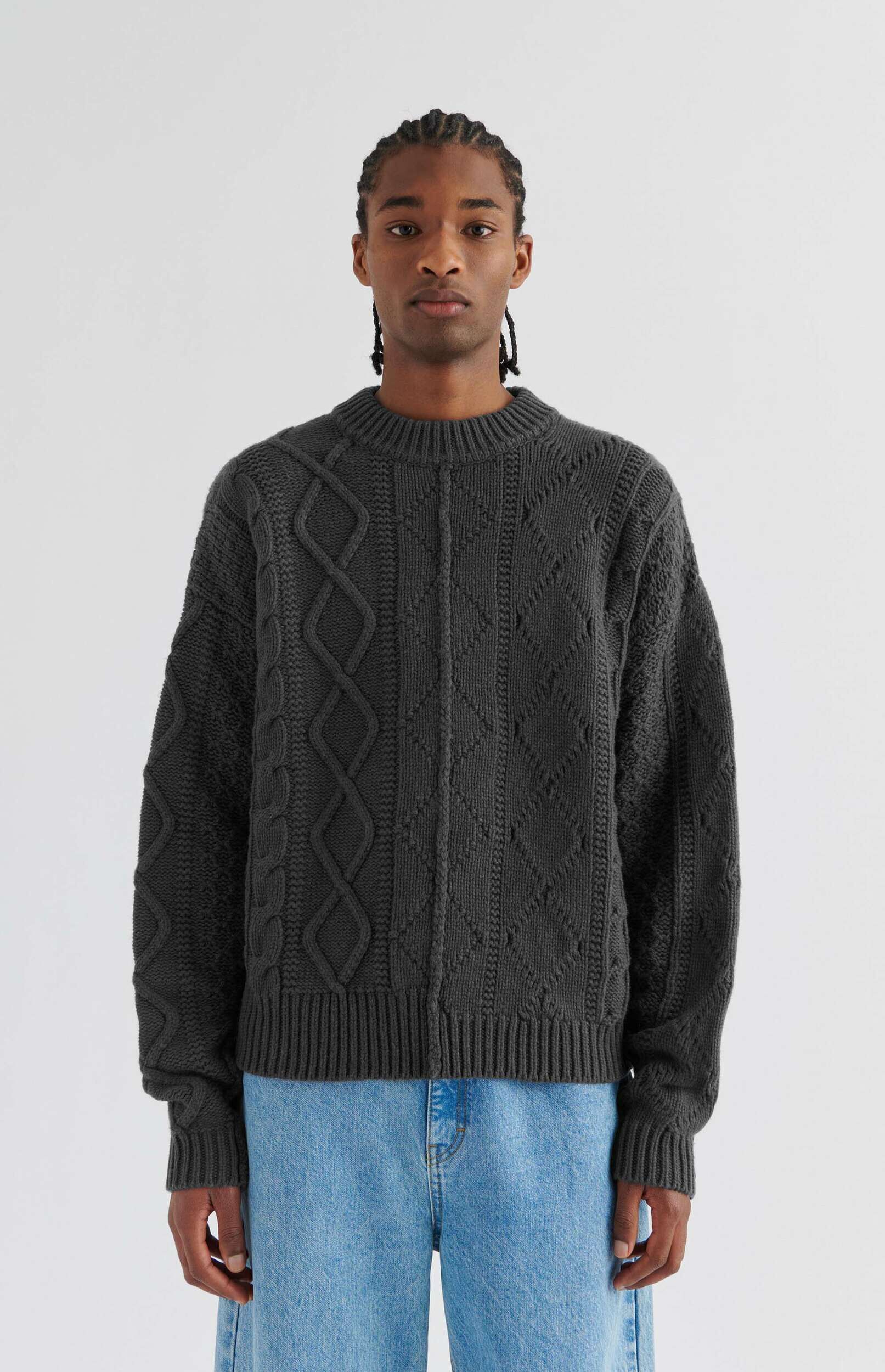 A man with cornrows wearing a dark grey cable-knit sweater with intricate patterns and light blue jeans, standing against a white background.