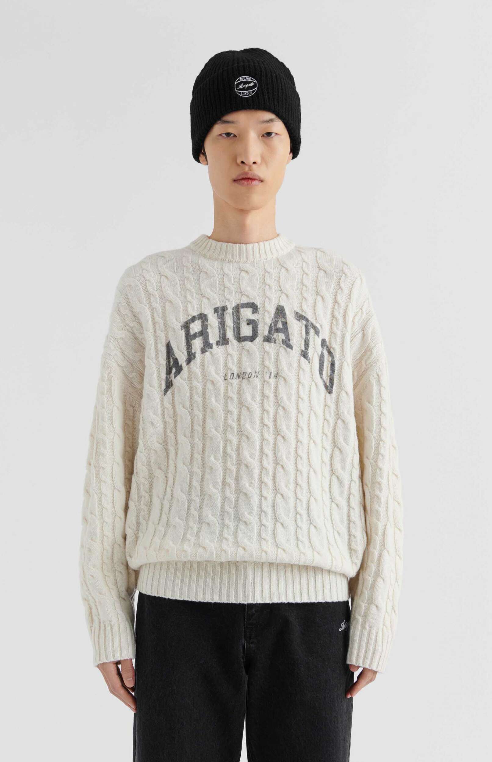A person wearing a cream cable-knit sweater with 'ARIGATO LONDON '14' text and a black beanie, standing against a white background.