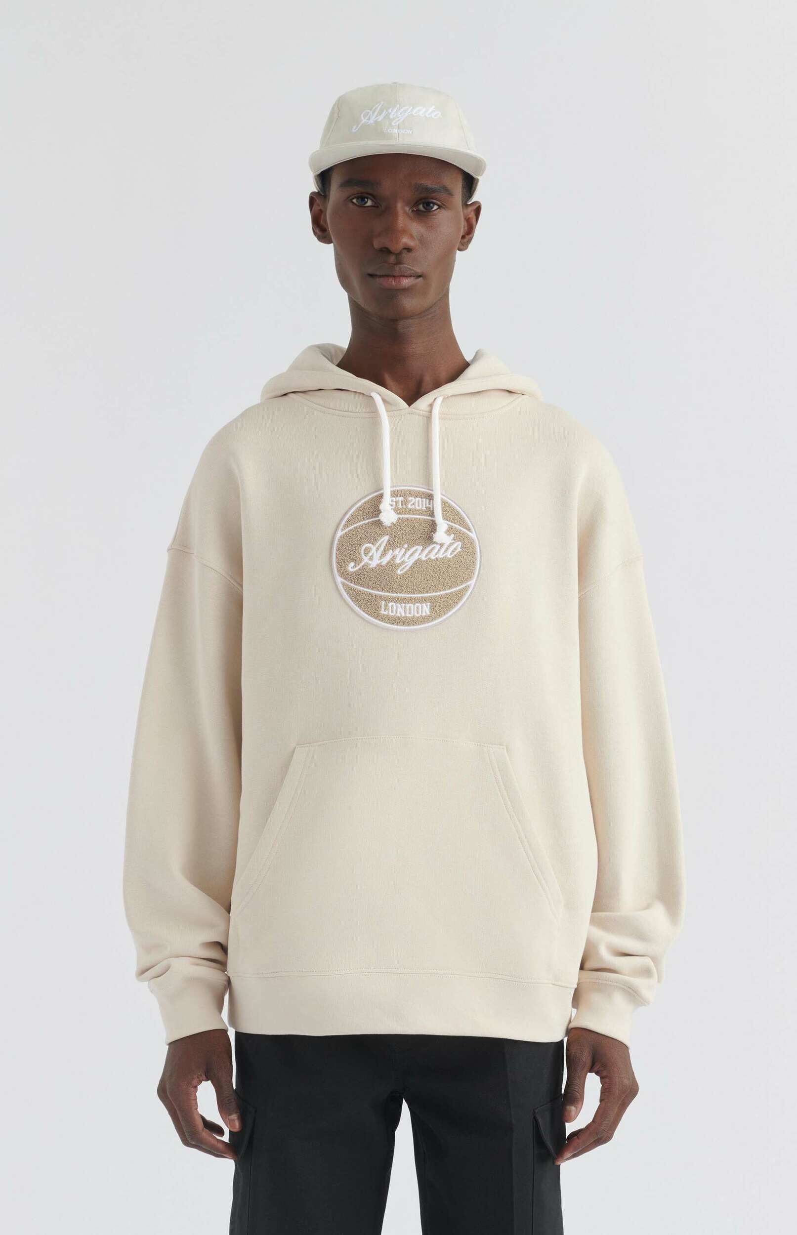 A man wearing a light beige hoodie with an 'Arigato LONDON' basketball-themed patch, black cargo pants, and a matching beige baseball cap, standing against a plain white background.