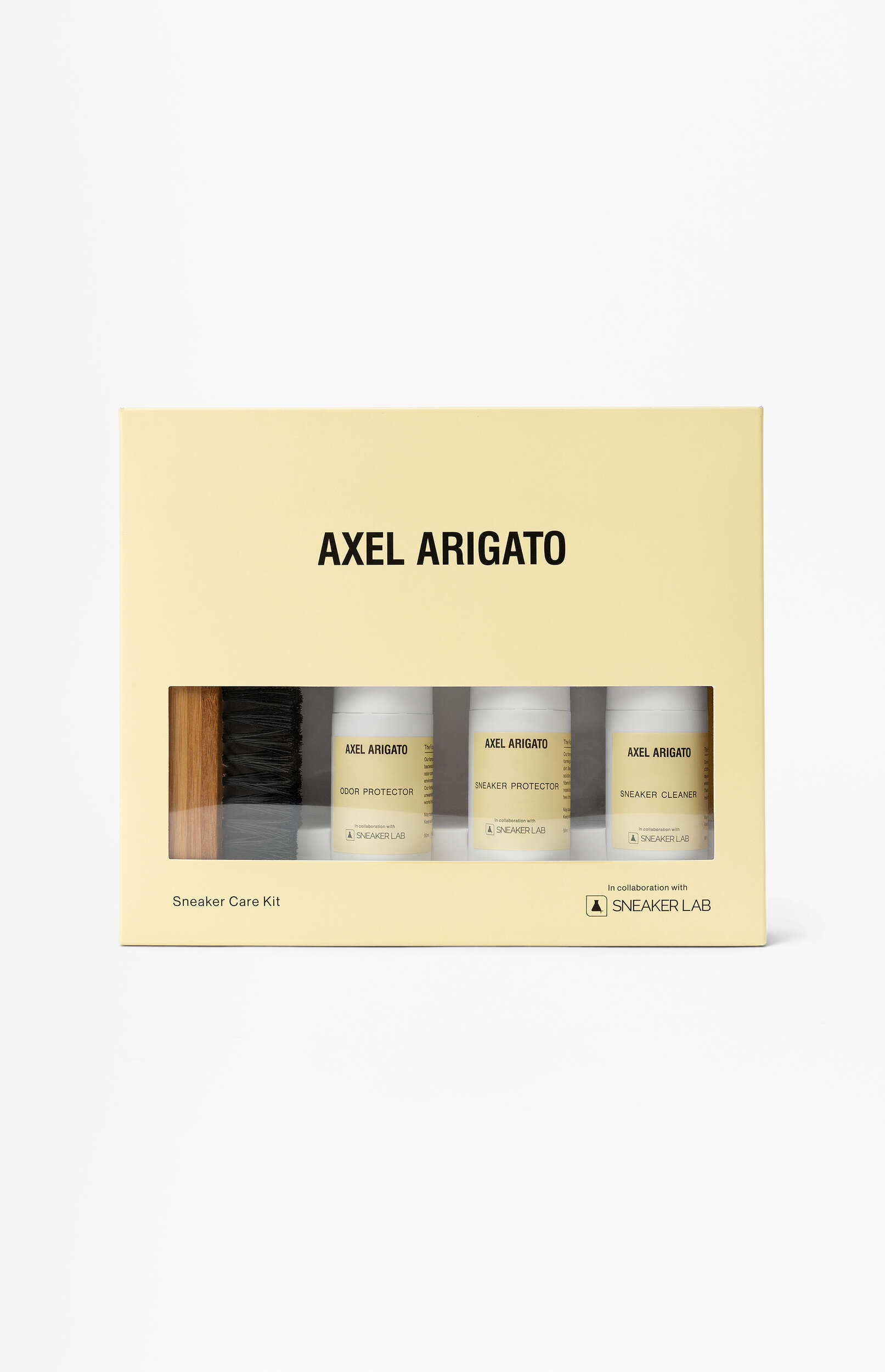 A light yellow box for an Axel Arigato Sneaker Care Kit, in collaboration with Sneaker Lab, displayed against a white background. The box has a clear window revealing a wooden-handled brush with black bristles and three white bottles labeled 'Odor Protector', 'Sneaker Protector', and 'Sneaker Cleaner'.