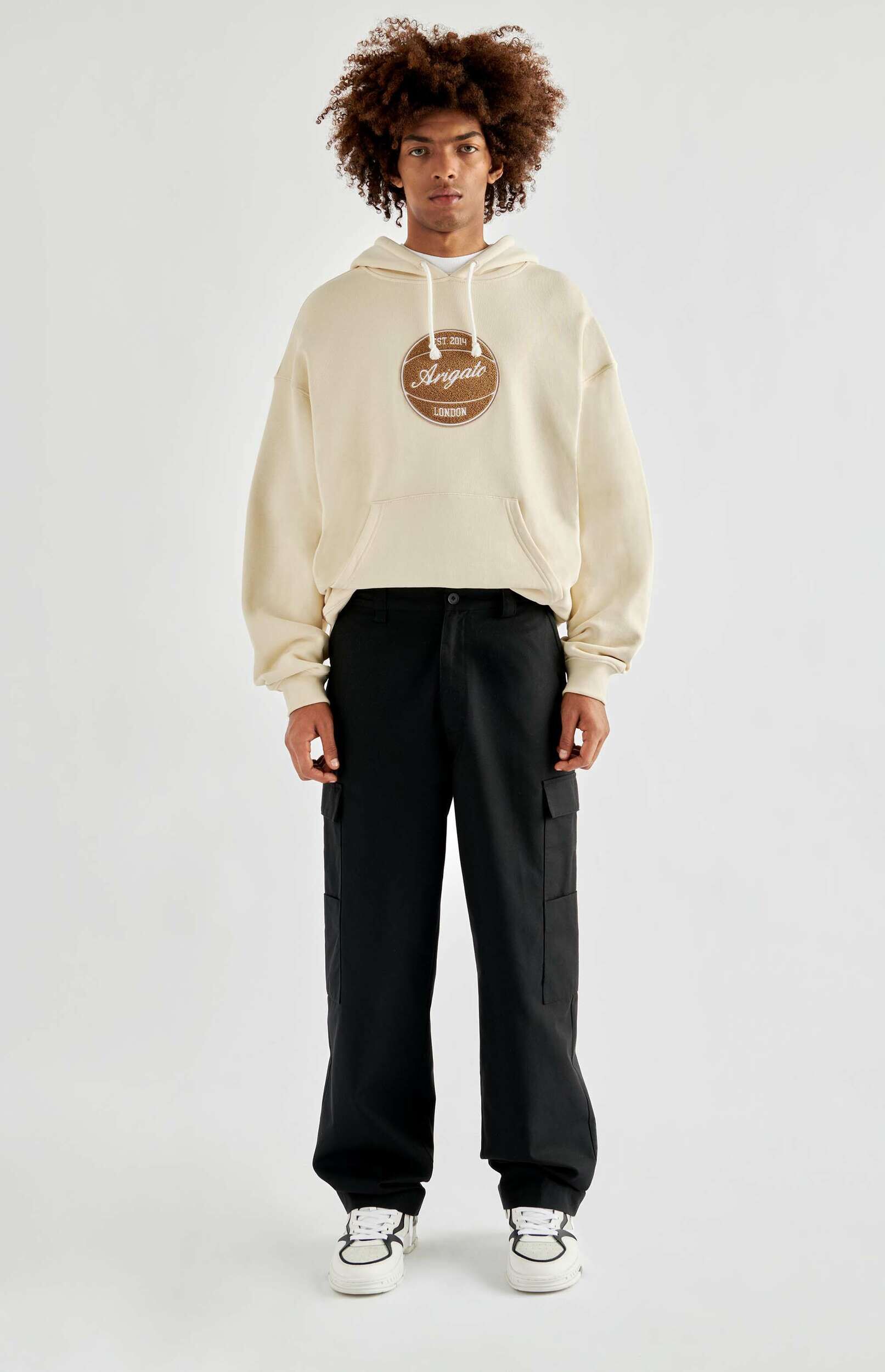 A man with curly brown hair stands full-body against a white background, wearing a cream-colored hoodie with a brown 'Arigato London EST 2014' logo patch, black wide-leg cargo pants, and white sneakers with black accents.