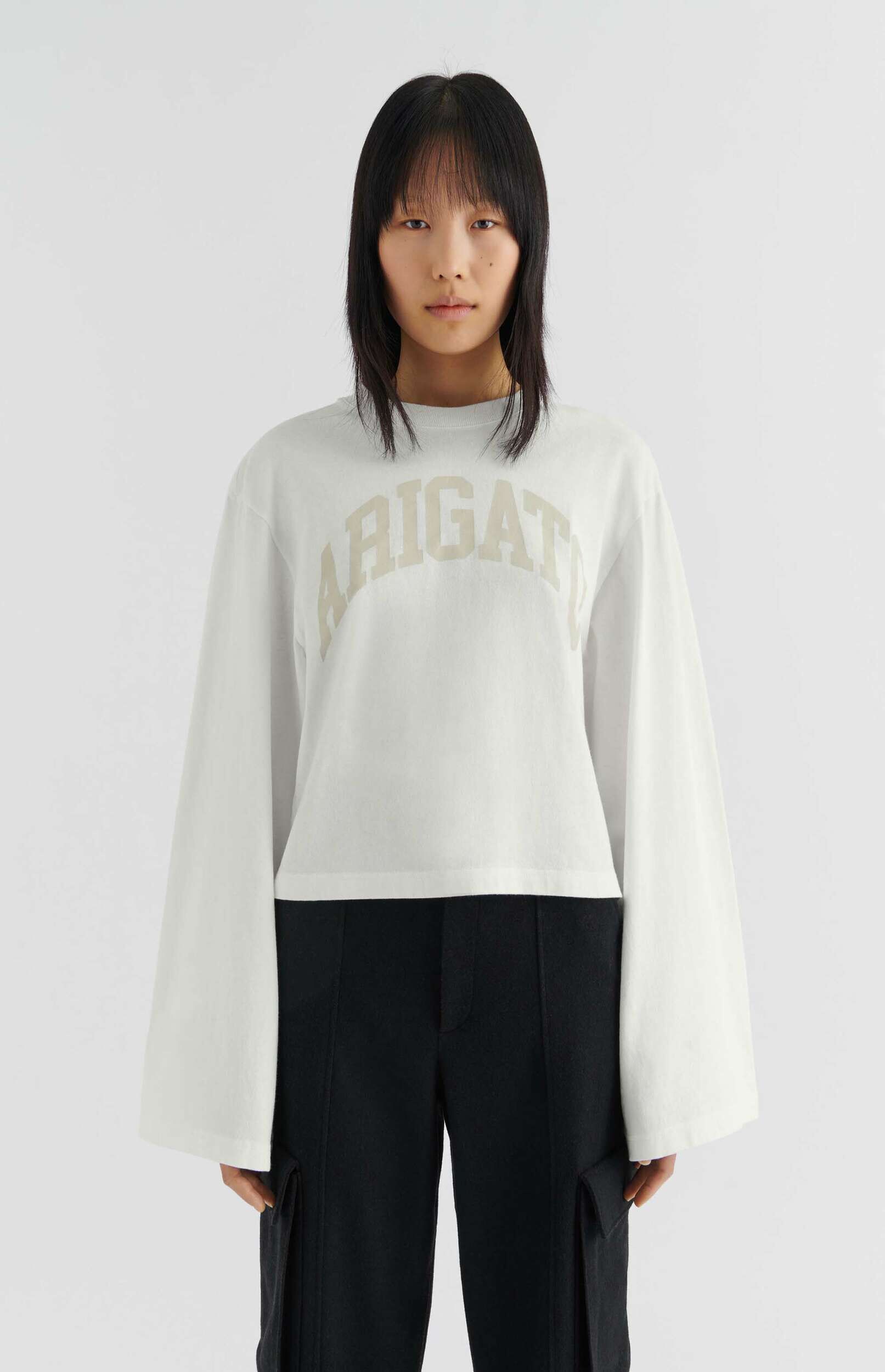 A person with straight black hair and bangs poses against a plain white background, wearing a loose white long-sleeve shirt with bell sleeves and a light beige 'ARIGAT' logo on the chest, paired with black wide-leg cargo pants.
