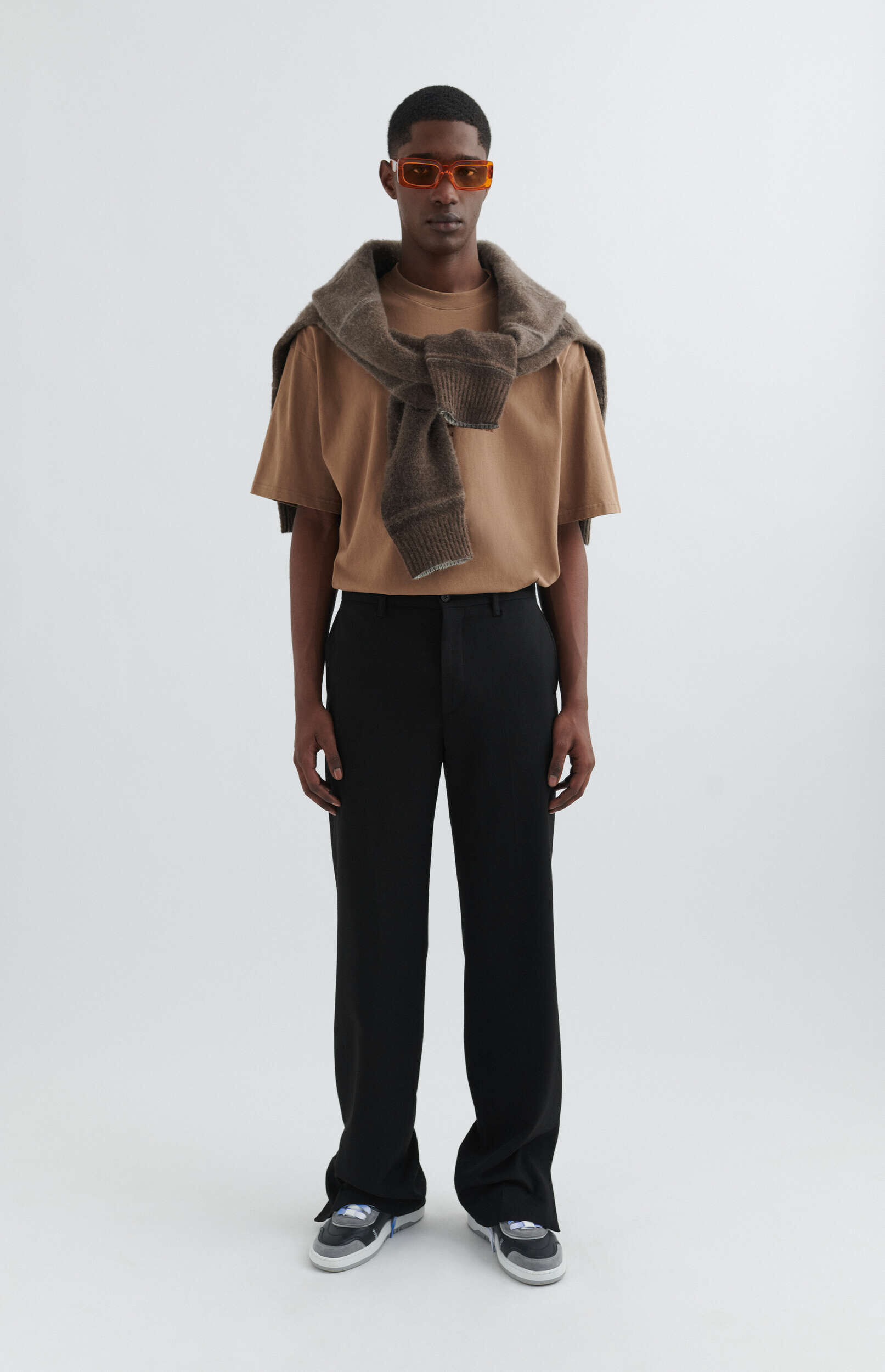 A full-length photo of a male model with dark skin, wearing orange rectangular sunglasses, a light brown short-sleeved t-shirt, a dark brown knitted sweater draped over his shoulders, black wide-leg trousers, and multi-colored sneakers, standing against a plain white background.