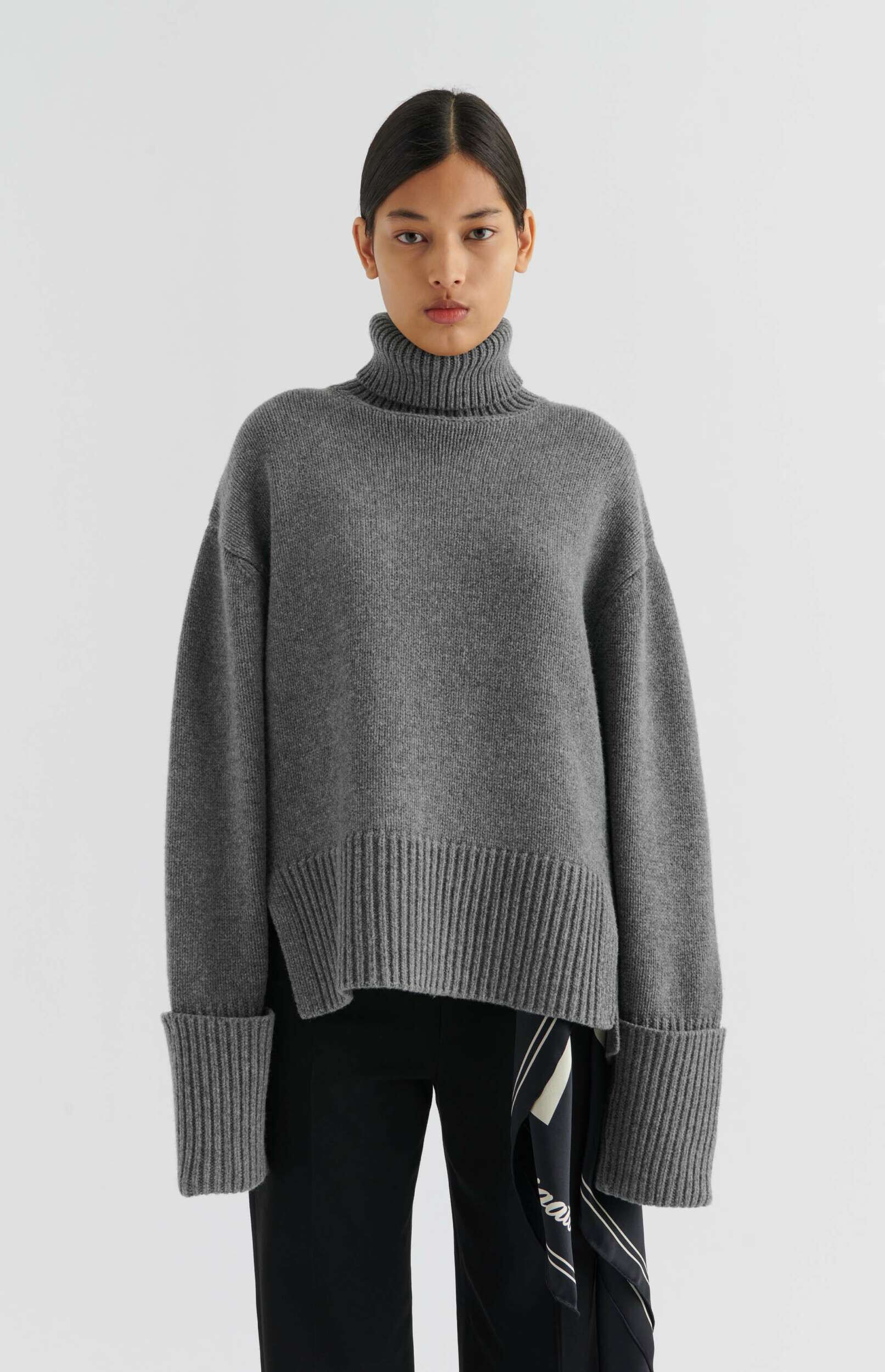A model stands front-facing, wearing an oversized, ribbed grey turtleneck sweater with wide cuffs and black trousers. Her dark hair is pulled back, and she has a neutral expression. A black and white patterned scarf is visible tucked into the trousers on her right side. The background is white.