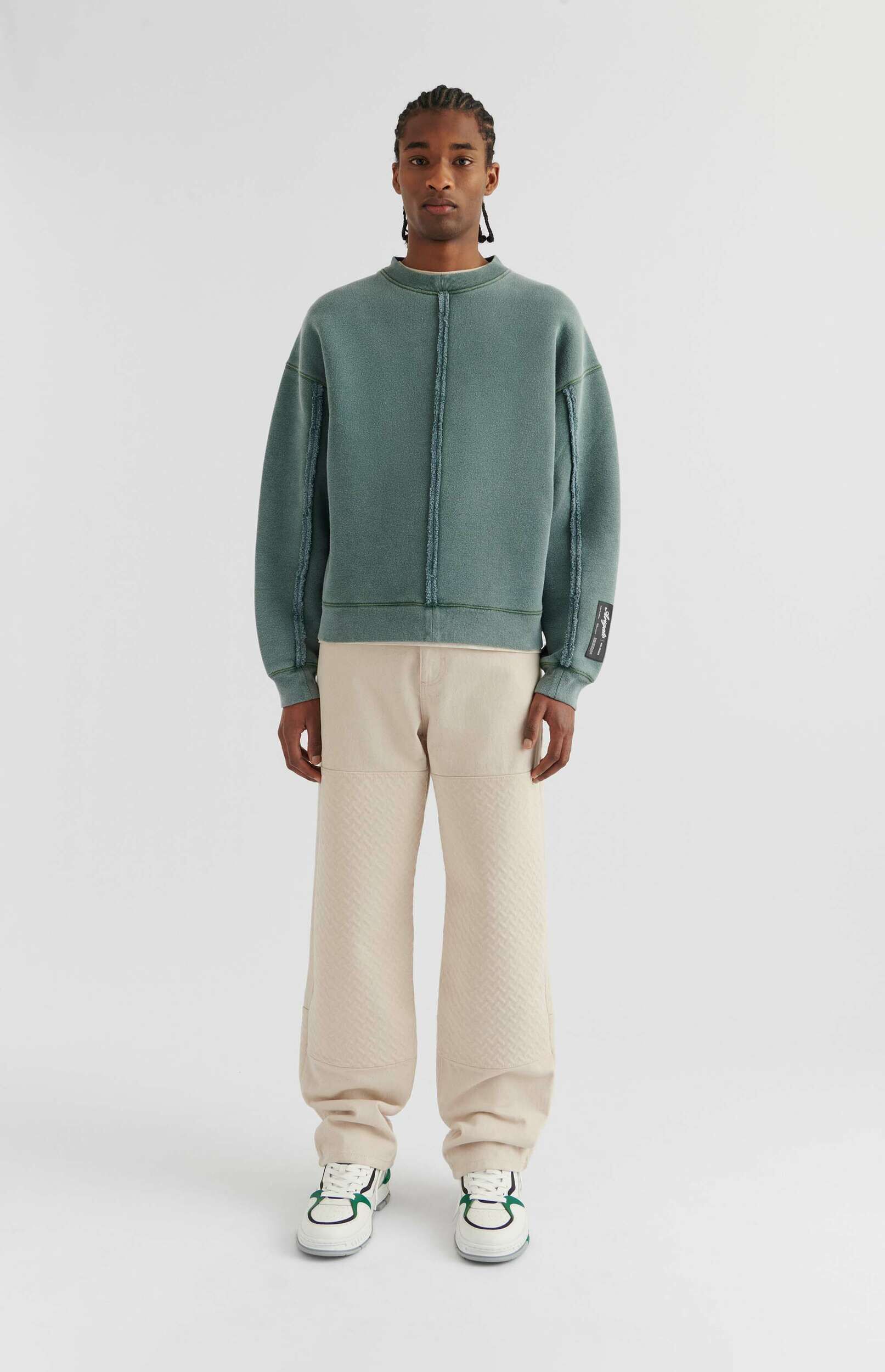 A full-body shot of a man wearing a teal oversized crewneck sweatshirt with visible seam details, light beige wide-leg pants with a subtle textured pattern, and white sneakers with green accents, standing against a plain white background.