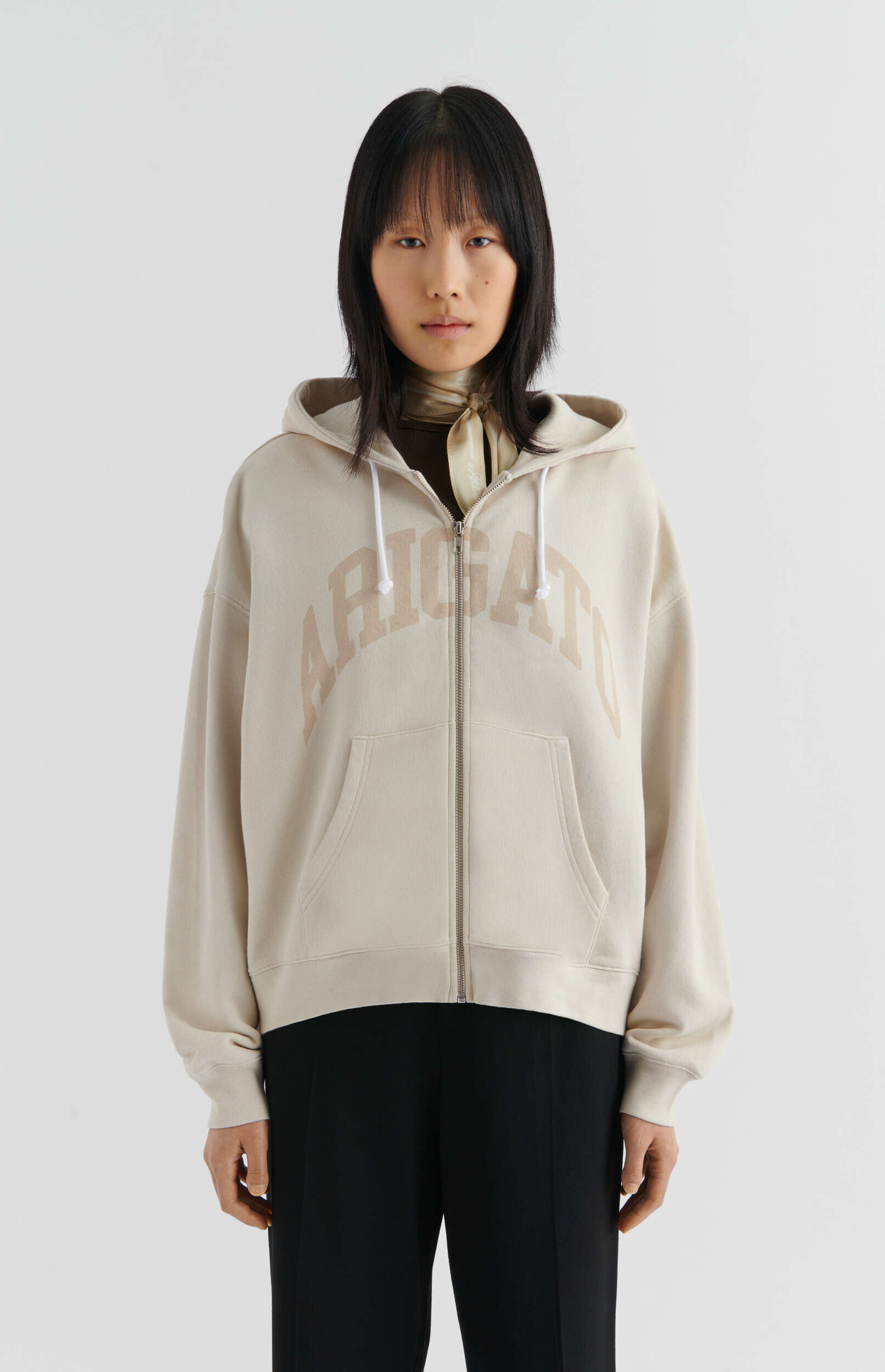 A person with straight black hair and bangs stands against a white background, looking directly at the viewer. They are wearing a light beige zip-up hoodie with 'ARIGATO' in darker beige letters across the chest, and white drawstrings. Underneath, a dark brown top and a light patterned scarf tied neatly around the neck are visible. The person is also wearing black trousers. The full-body shot is from the waist up.