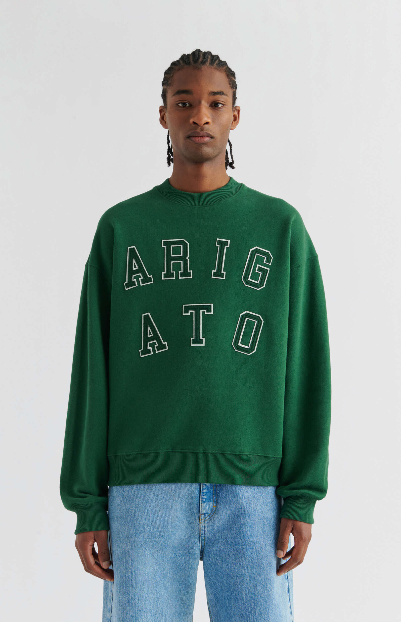 A young man with braided hair stands against a plain white background, wearing a dark green crew-neck sweatshirt with the word 'ARIGATO' in white outlined letters across the chest, and light blue relaxed-fit jeans.