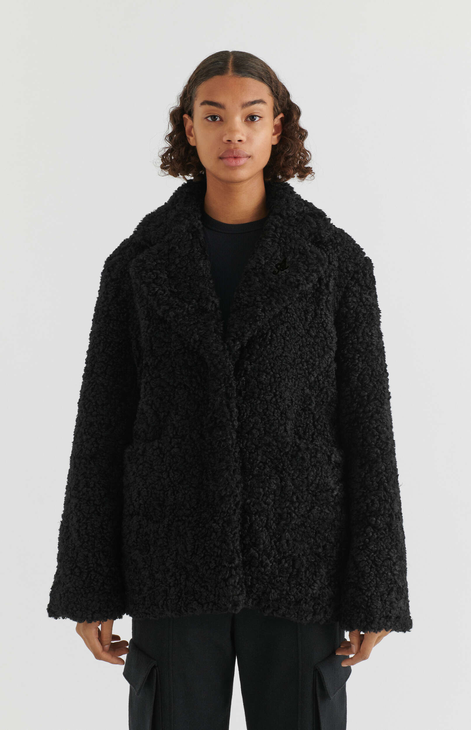 A young woman with curly brown hair poses wearing an oversized black textured boucle coat over a black ribbed top and black cargo pants, standing against a clean white background.