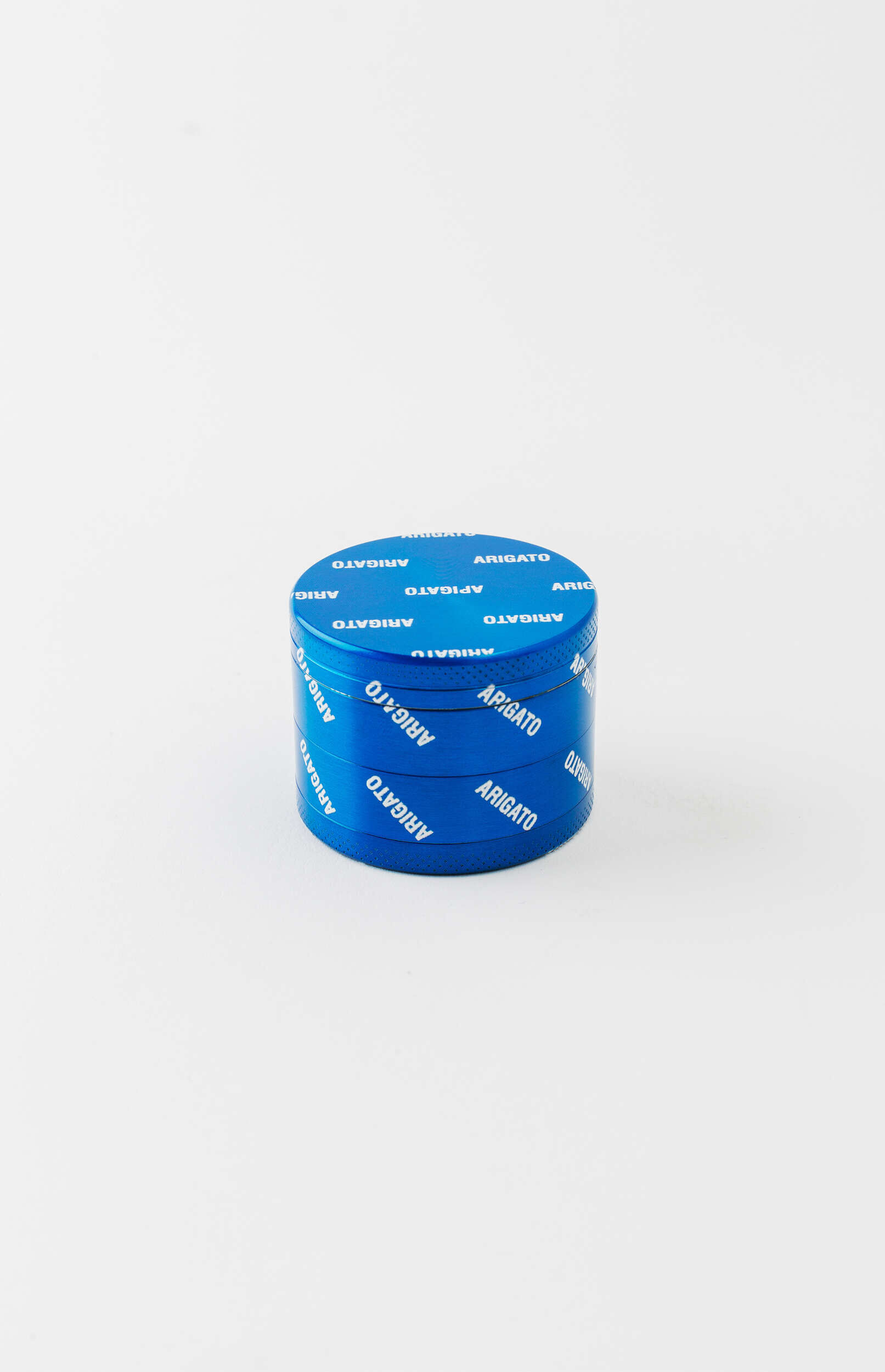 A blue, cylindrical herb grinder with the word 'ARIGATO' printed repeatedly in white letters around its entire surface. The grinder has a textured bottom edge and is positioned upright on a clean white background.