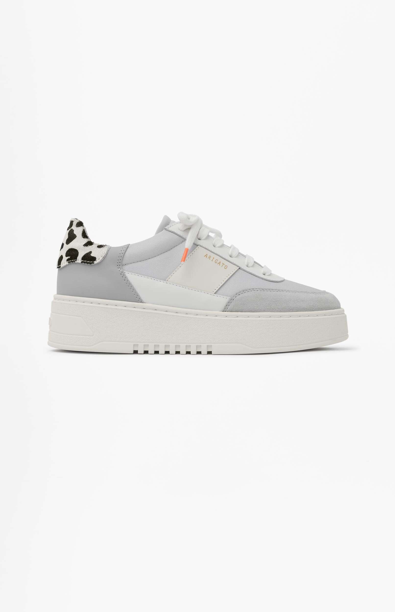A light gray and white sneaker with a chunky white sole and a black and white spotted animal print detail on the heel. The brand 'ARIGATO' is visible in gold letters on the side.
