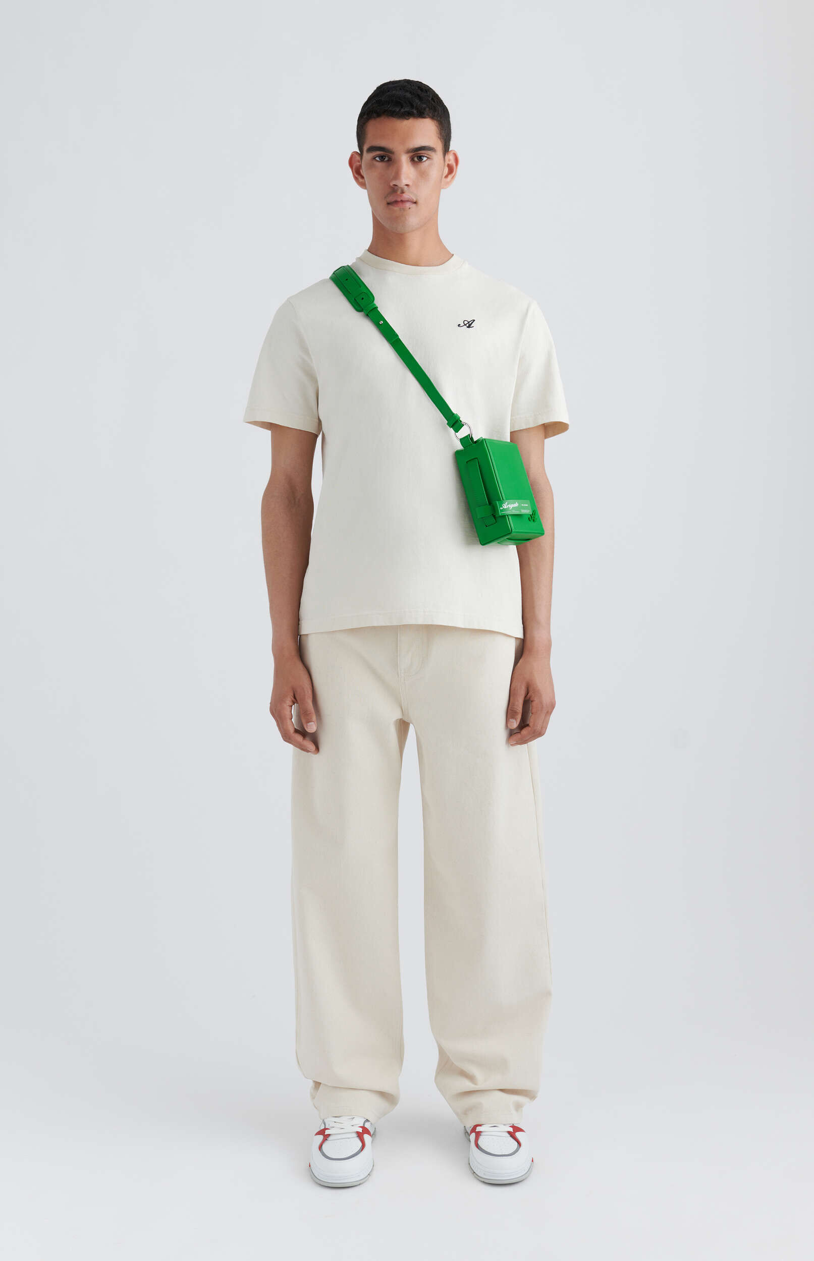 A male model stands full length against a white background, wearing an off-white t-shirt with a small dark logo and matching wide-leg trousers, complemented by a bright green rectangular cross-body bag and white sneakers with red and grey details.
