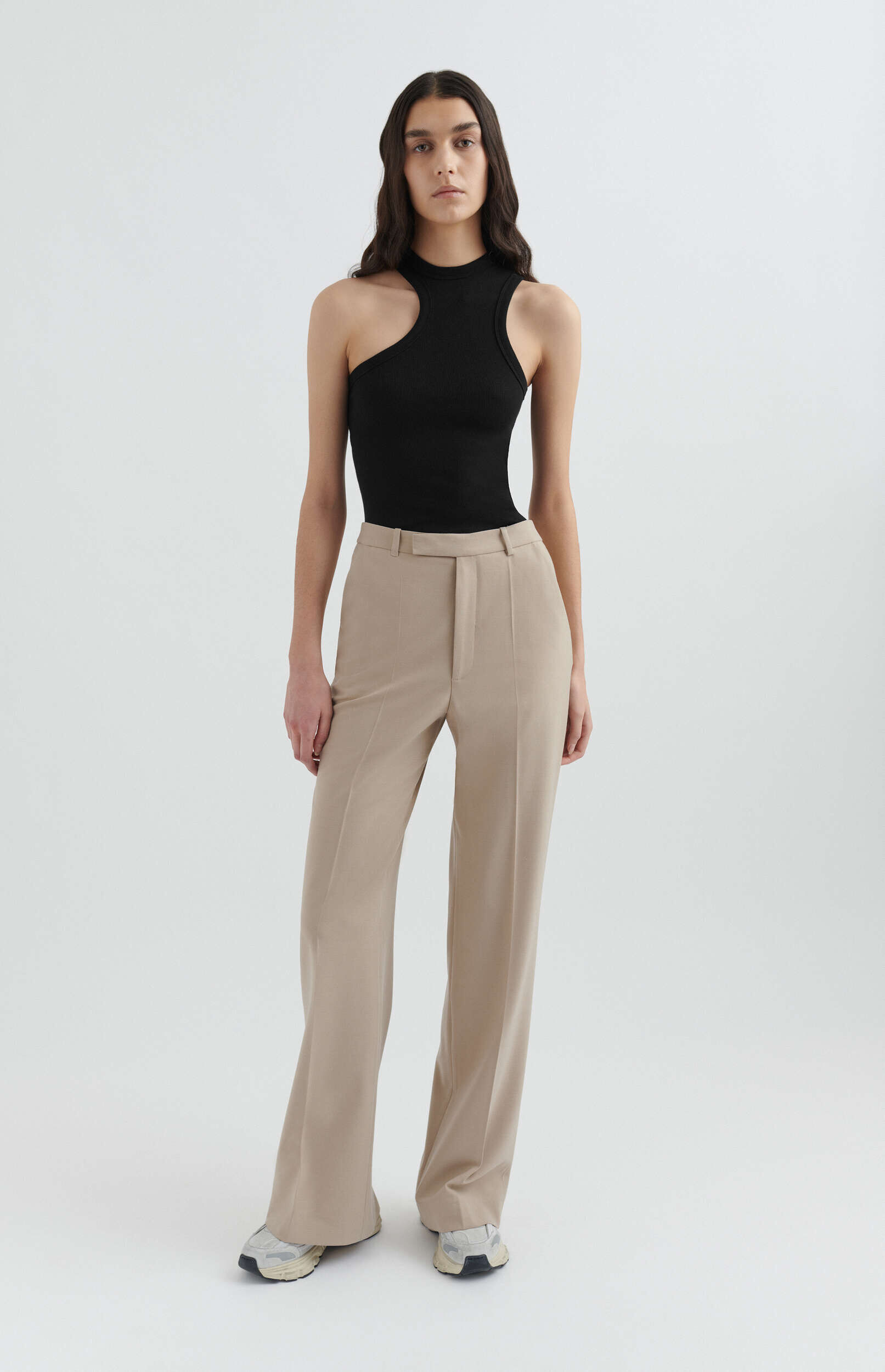 A woman with long dark hair models a black ribbed one-shoulder top and high-waisted, wide-leg beige trousers, paired with light sneakers, standing against a plain white background.
