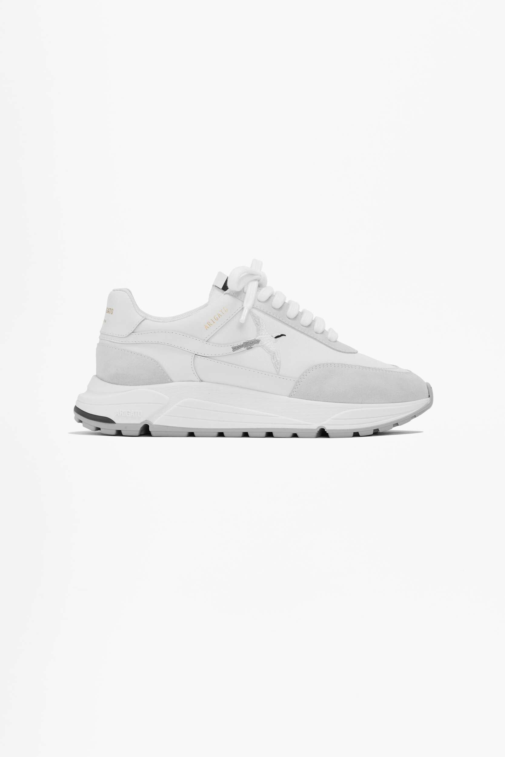 A white Axel Arigato Genesis vintage runner sneaker is shown at an angle against a plain white background. The shoe features white leather and light grey suede panels, white laces, a chunky white sole with grey tread, and the 'ARIGATO' branding in gold on the side with a bird emblem.