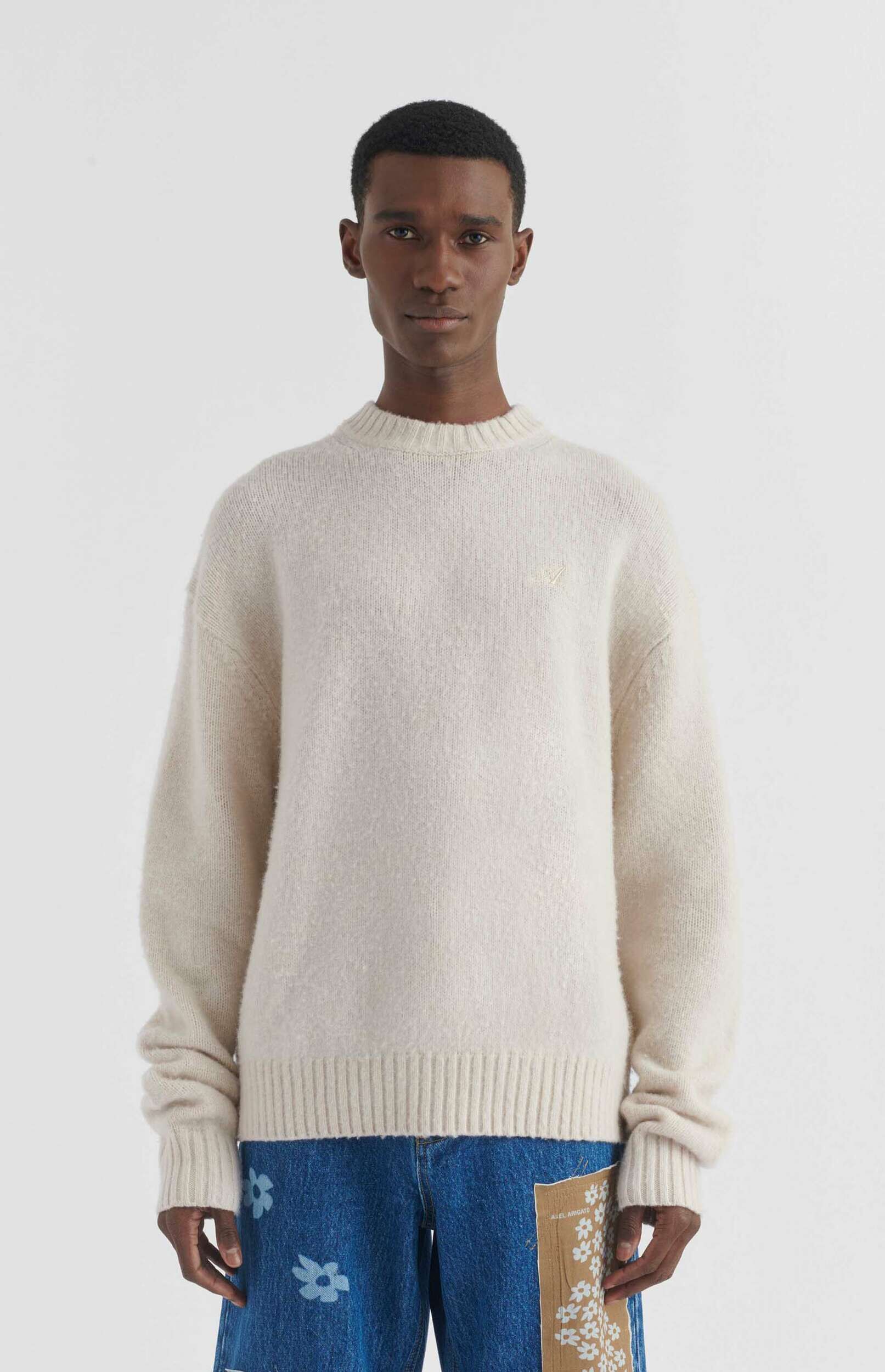 A young man, medium skin tone, with short dark hair, stands facing forward against a plain white background. He is wearing a cozy, light cream or off-white crew-neck sweater with a subtle, light-colored embroidered logo on the left chest. Below the sweater, he wears blue denim jeans featuring light blue stylized flower patterns and a large, light brown patch on the right thigh with smaller white flower patterns and the text 'AXEL ARIGATO'.