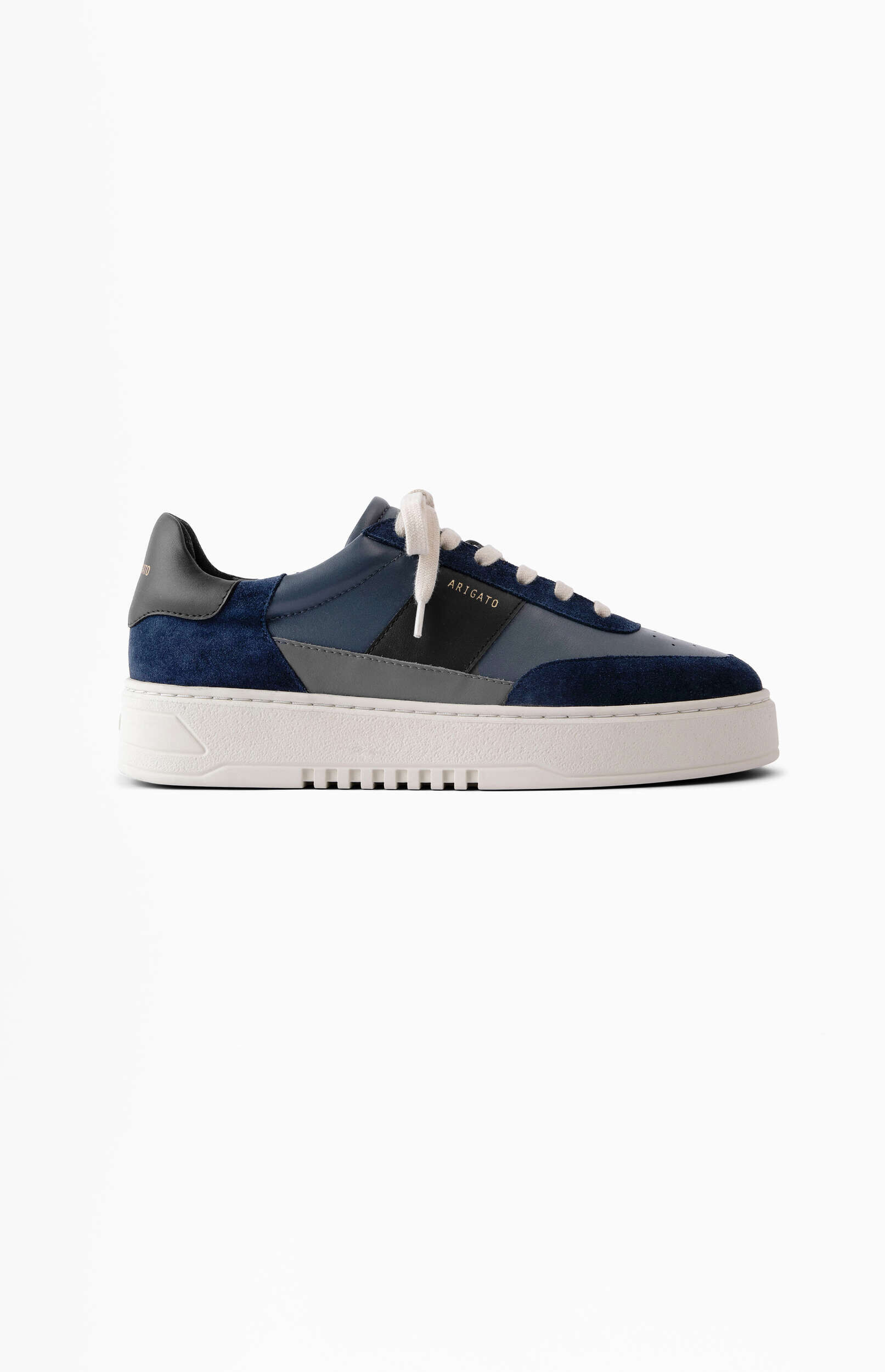 A side view of a men's low-top sneaker, featuring a design with navy blue suede, medium blue leather, and gray leather panels. A black stripe on the side displays the gold 'ARIGATO' logo. The shoe has white laces and a thick white sole with a textured tread pattern.