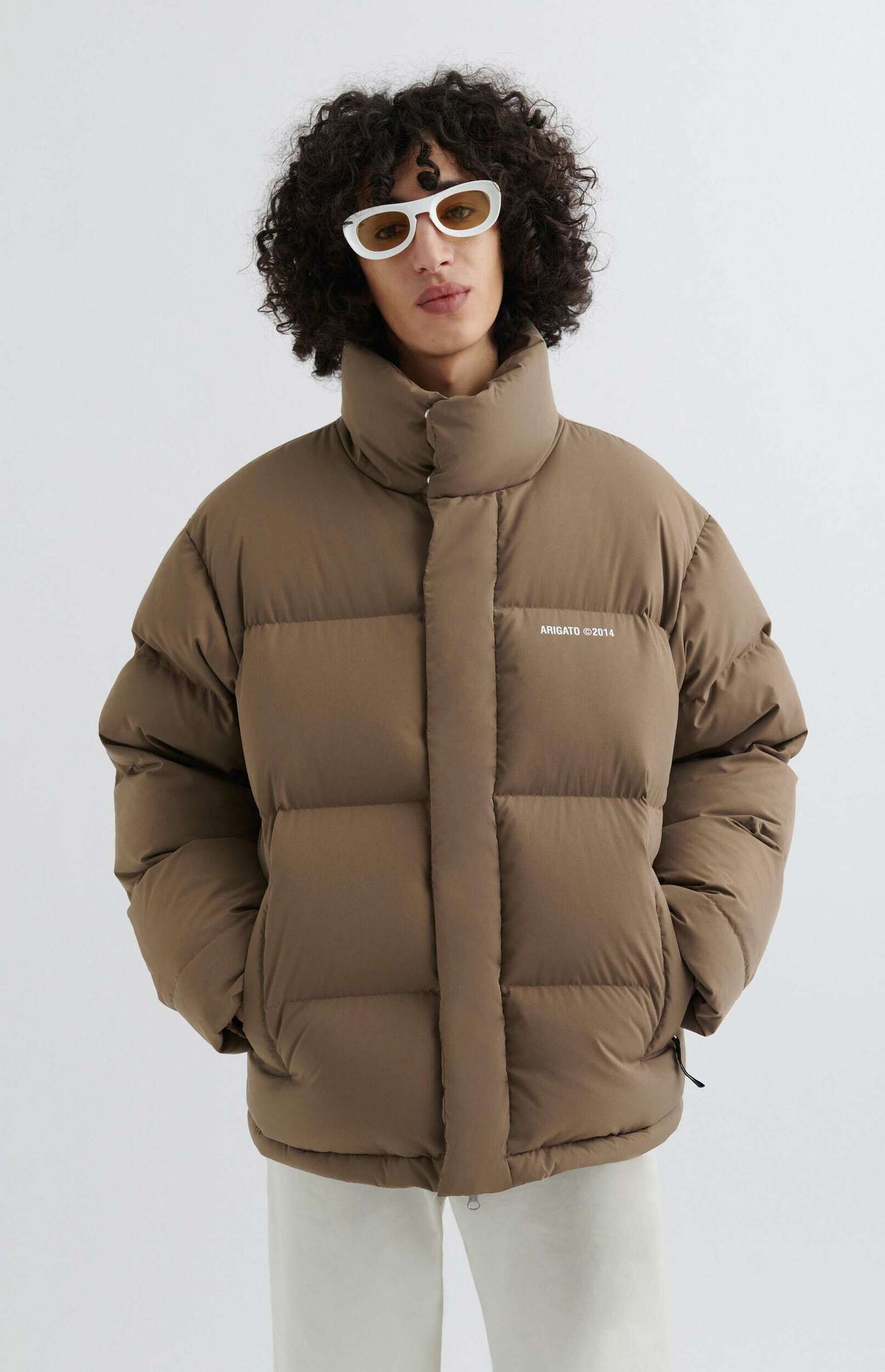 A person with curly dark hair wearing a brown puffer jacket with 'ARIGATO ©2014' printed on the chest, and white retro sunglasses with amber lenses, standing against a plain white background.