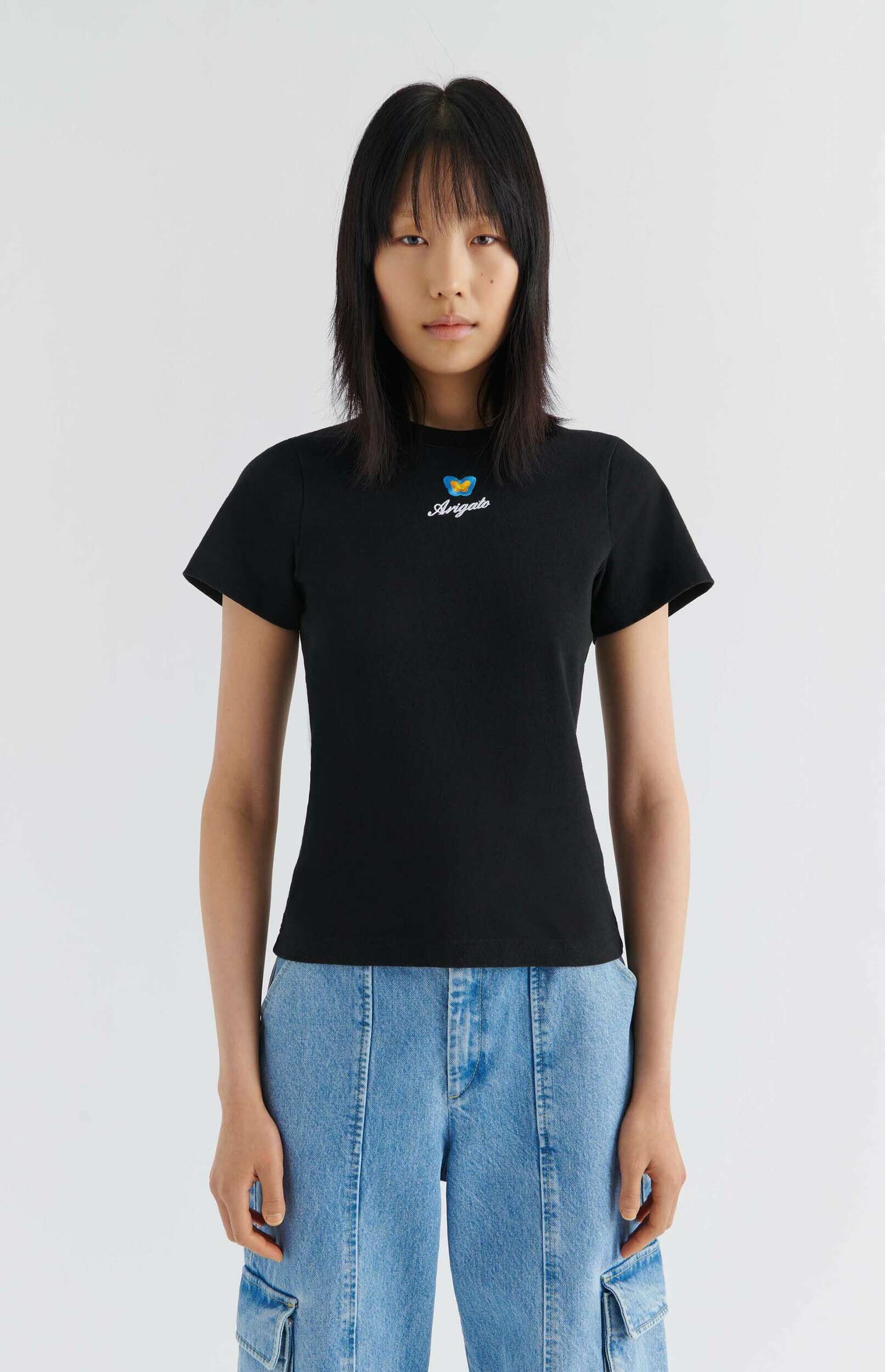 A young woman with dark, straight hair and bangs stands against a white background, wearing a black t-shirt with a blue and yellow butterfly graphic and the word 'Angels' in script, paired with light blue wide-leg denim cargo pants.