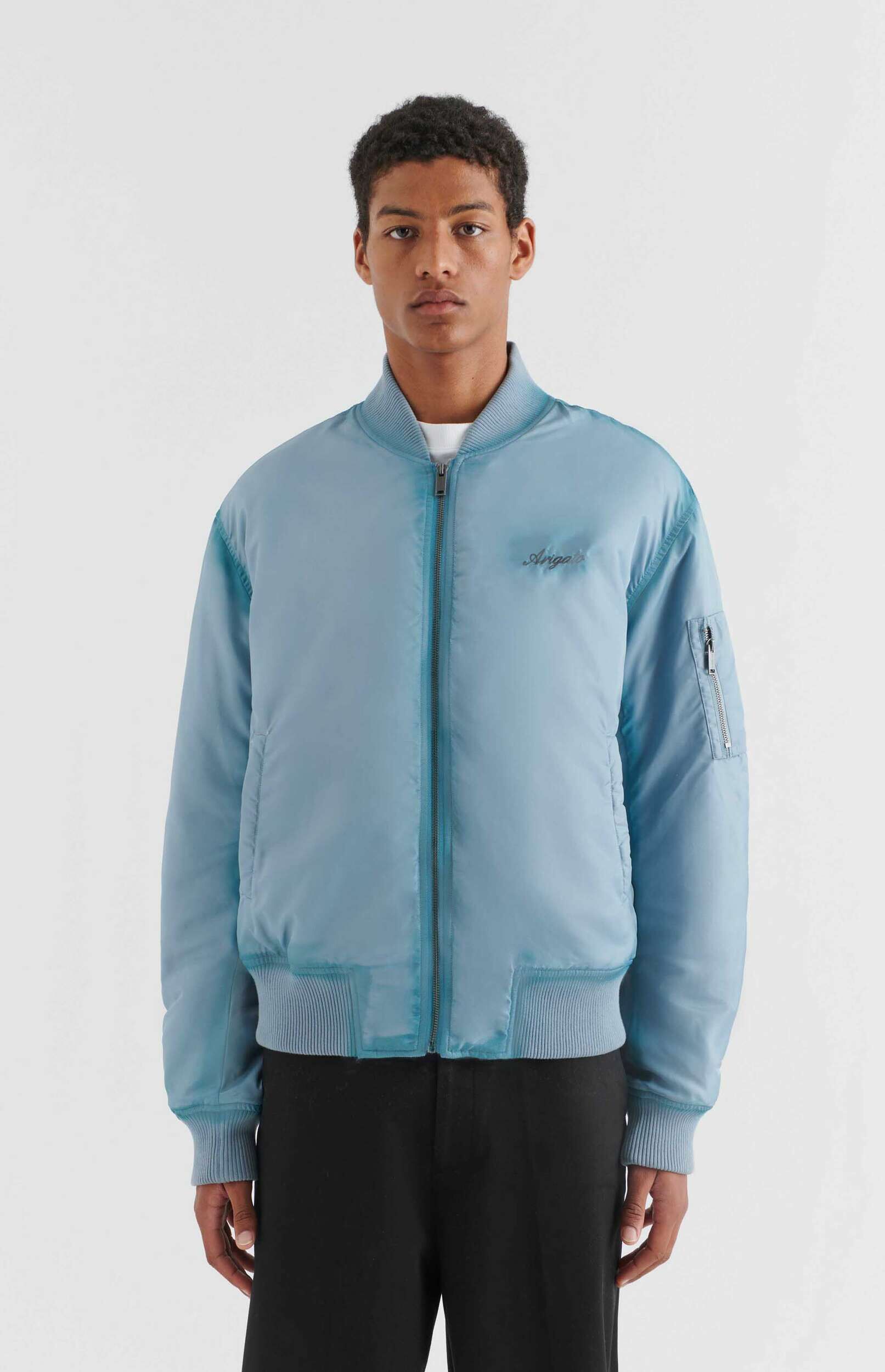 A young man with dark, curly hair stands against a white background, wearing a light blue bomber jacket with a full zipper, ribbed collar, cuffs, and hem, and a subtle 'Arigato' logo on the left chest. He is also wearing black trousers.