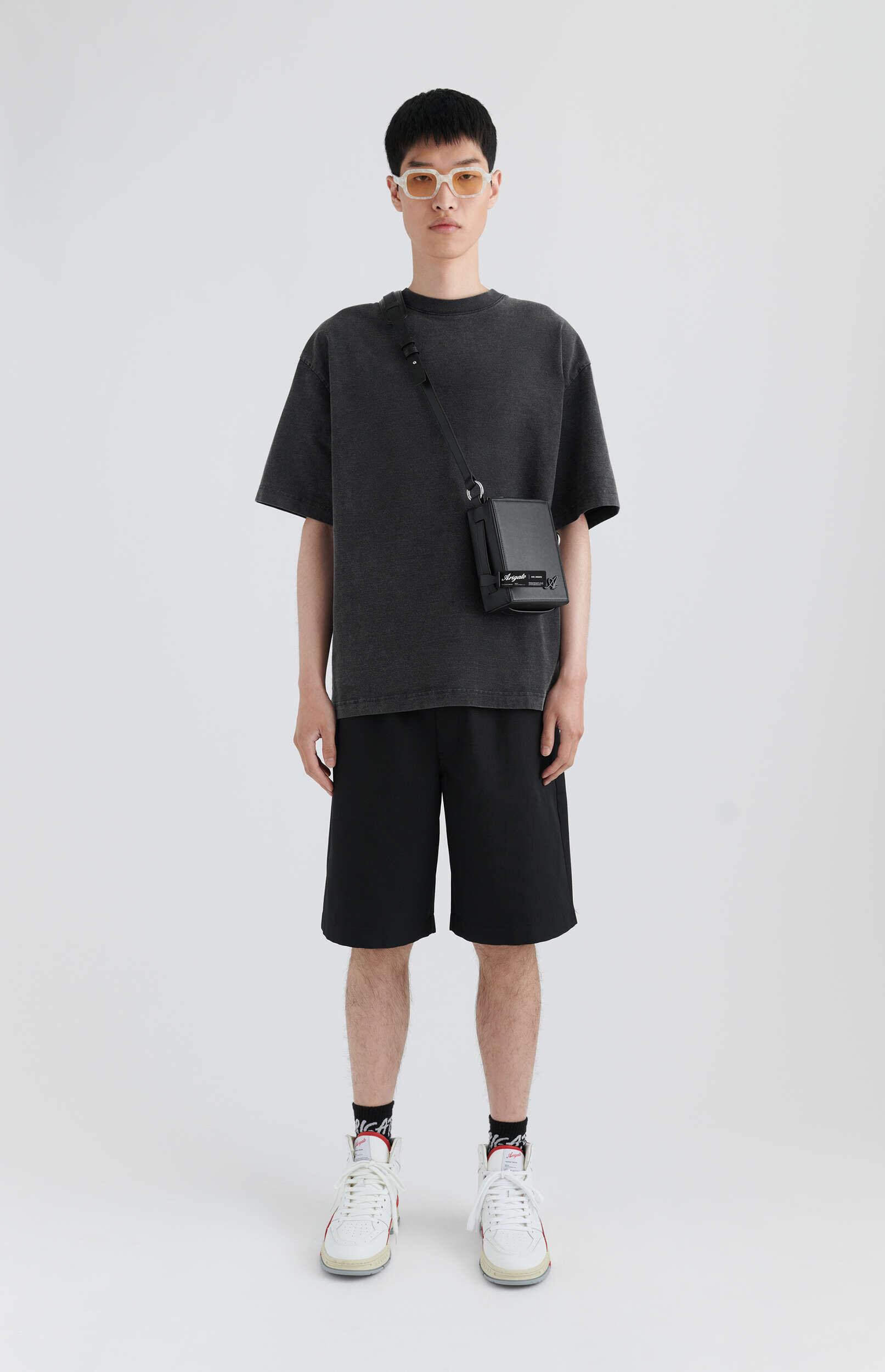 A young man of East Asian descent stands full-length against a white background, dressed in a washed black oversized t-shirt, black athletic shorts, and white high-top sneakers with red accents. He wears clear-framed, orange-tinted rectangular sunglasses and a black crossbody bag with a white logo.
