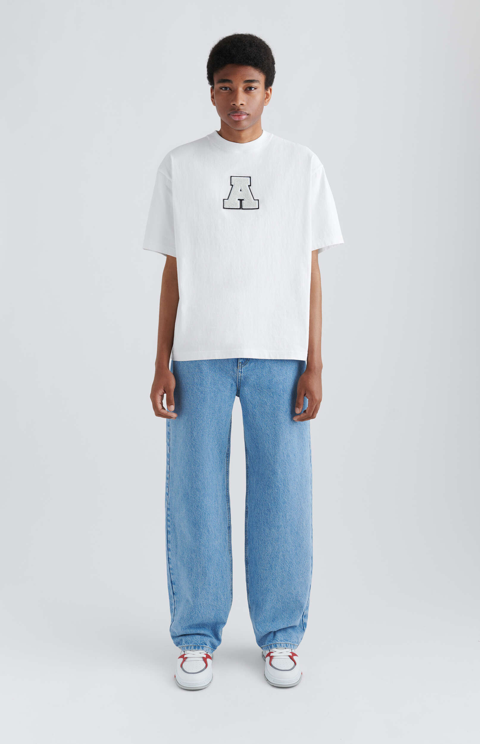 A young person with dark skin and short dark hair stands centered against a plain white background. They are wearing a white short-sleeved t-shirt featuring a large, stylized letter 'A' with a black outline and light gray fill on the chest, paired with light blue, loose-fitting denim jeans and white sneakers with red accents. The person has a neutral expression and their hands are visible at their sides.