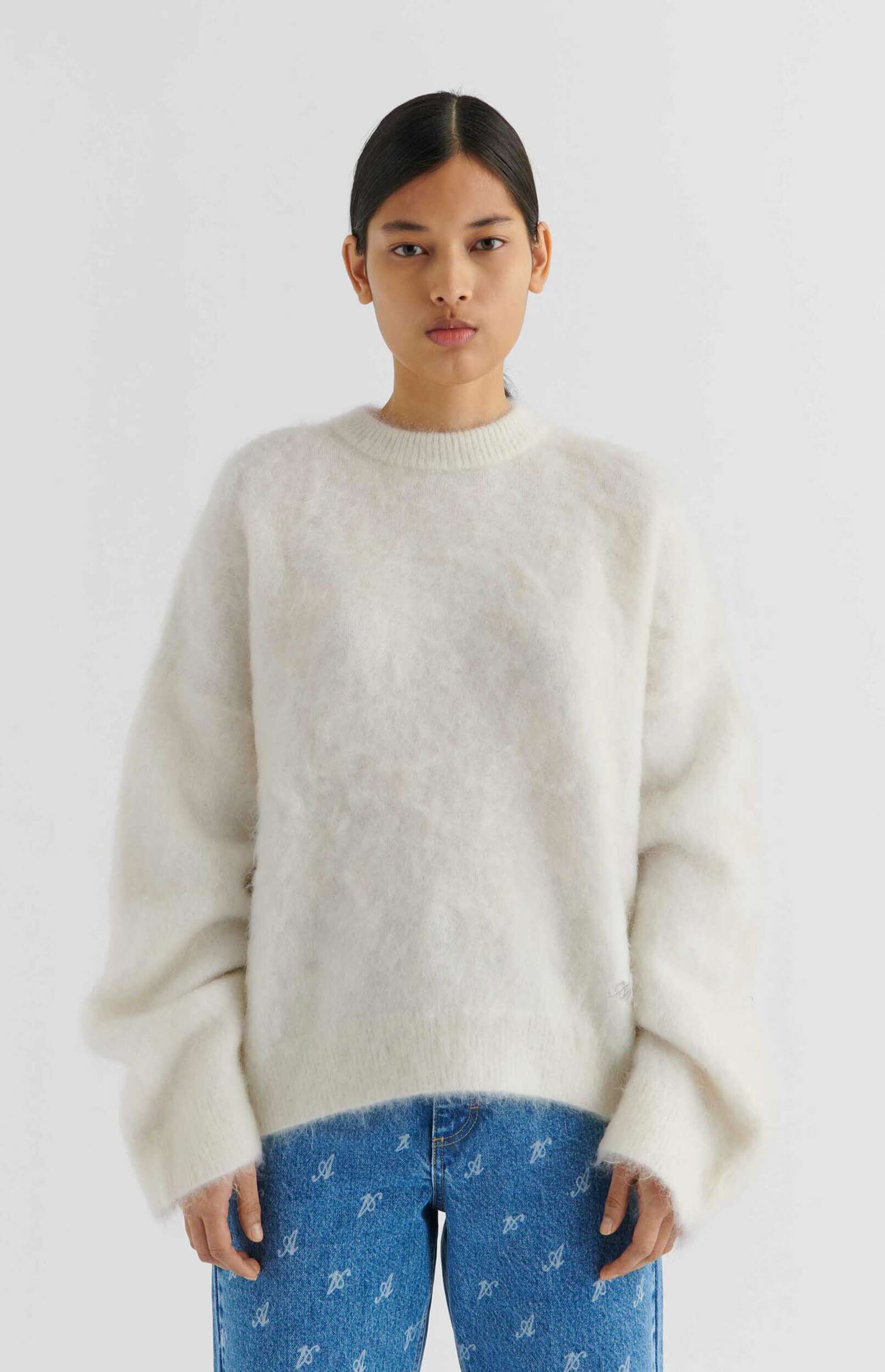 A woman wearing a fluffy white crew-neck sweater and blue jeans with a repeating white 'A' pattern stands against a plain white background.