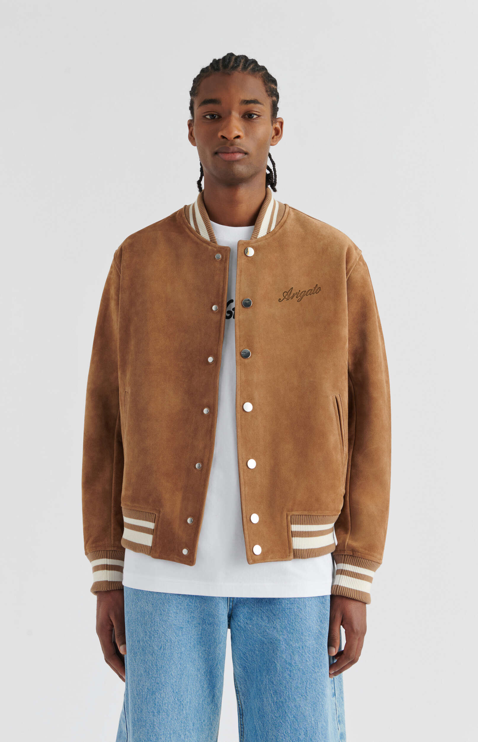 A person with braided hair stands against a white background, wearing a brown suede bomber jacket with white and brown striped ribbed collar, cuffs, and hem. The jacket has silver snap buttons and 'Arigato' embroidered in black cursive on the left chest. They are also wearing a white t-shirt and light blue jeans.