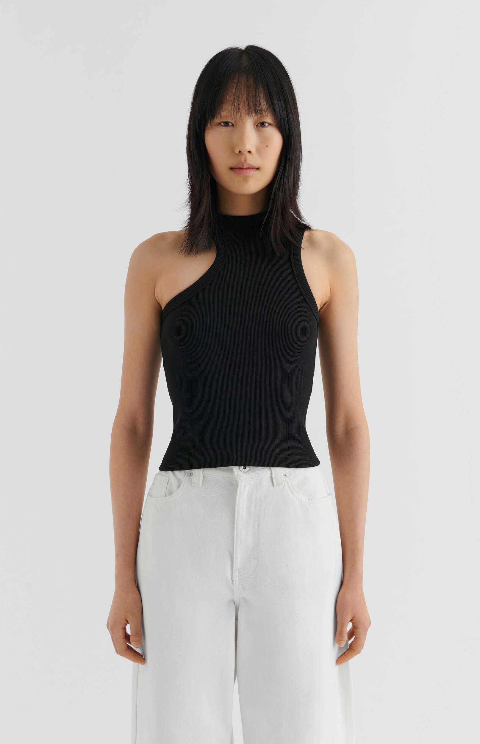 A young woman with dark hair wears a fitted black ribbed one-shoulder top and white wide-leg jeans, standing against a plain white background.