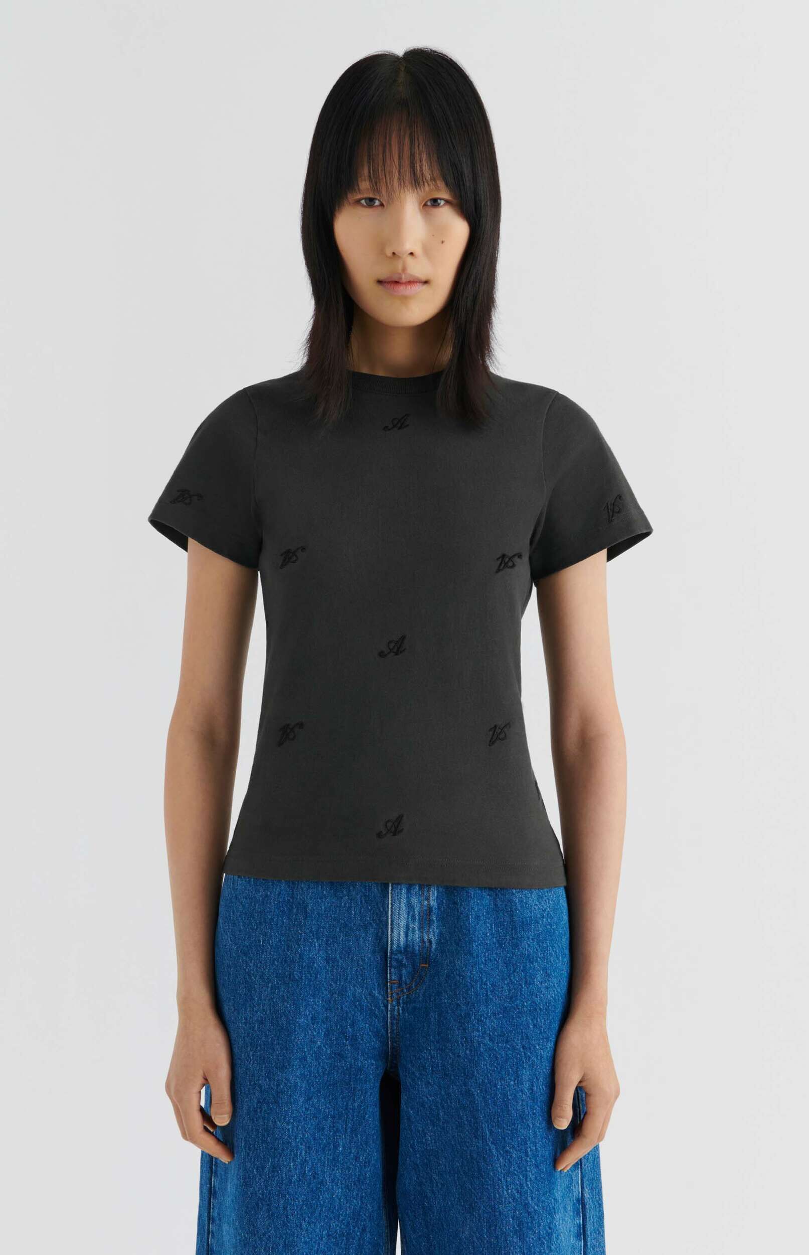 A person with straight dark hair and bangs stands against a white background, wearing a dark short-sleeve t-shirt with a subtle repeating pattern of embroidered 'JS' and 'A' letters, paired with blue denim jeans.
