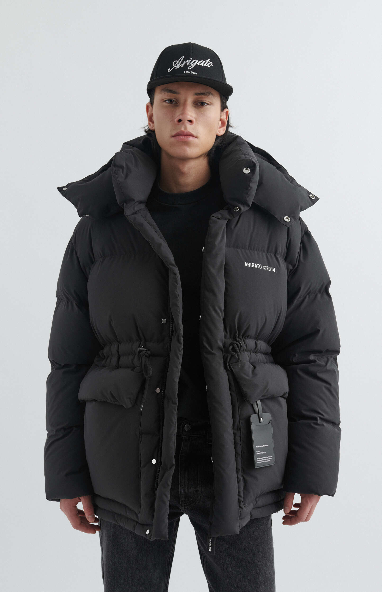 A man wearing a black puffer jacket with a drawstring waist and a black baseball cap with 'Arigato London' written on it, standing against a white background.