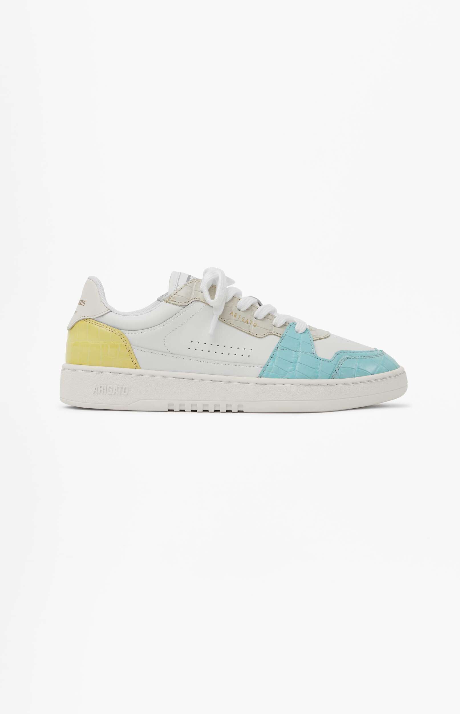 A white leather low-top sneaker with light yellow and light blue crocodile-embossed accents. The brand name 'ARIGATO' is visible on the side of the white sole. The shoe is positioned on a plain white background.