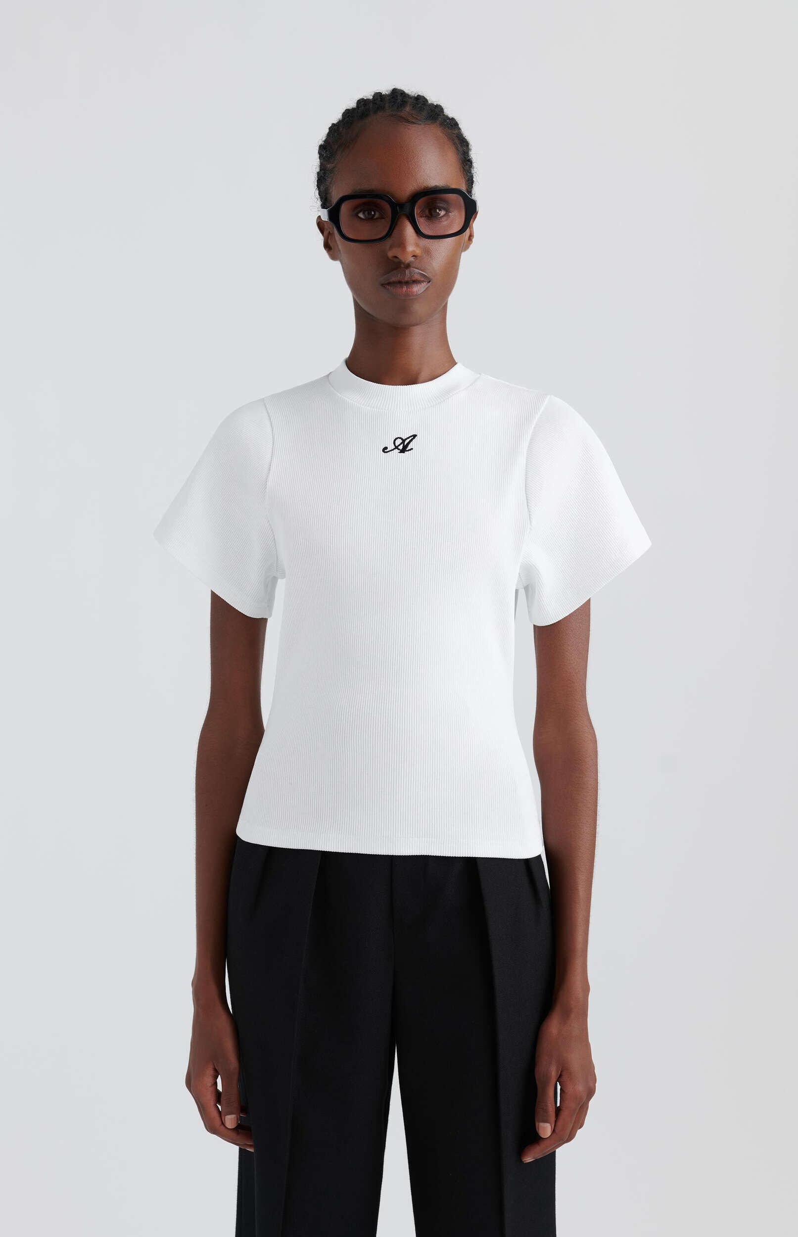 A person with dark skin and braided hair, wearing rectangular black-framed glasses, a white ribbed short-sleeve top with a black 'A' logo on the chest, and black wide-leg pants. They stand facing forward against a plain white background.