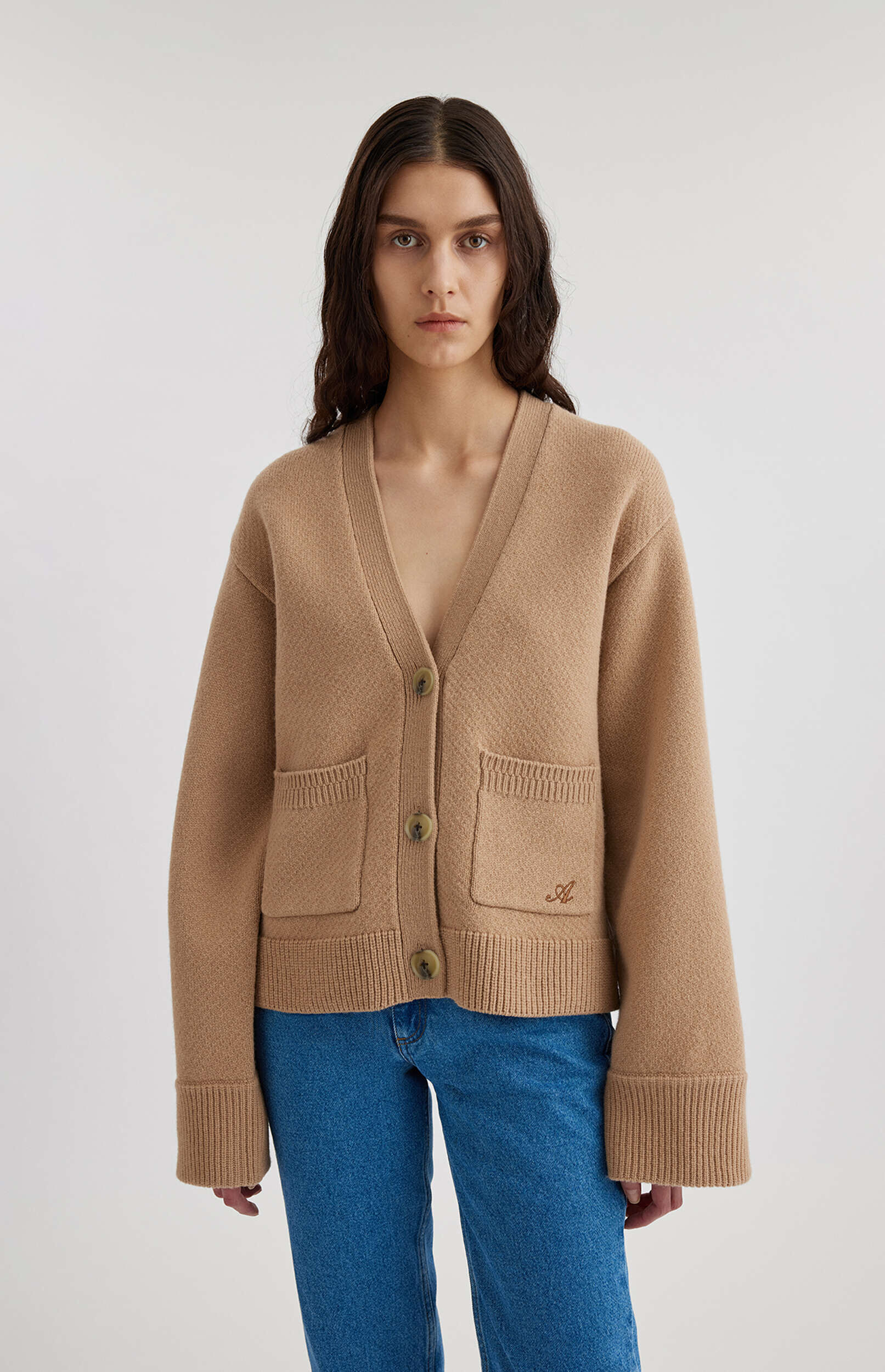 A full-length shot of a woman with long dark hair, wearing a camel-colored V-neck knit cardigan with three large brown buttons, two front patch pockets (one with a subtle embroidered 'A' initial), and ribbed cuffs and hem, paired with blue jeans, against a plain white background.
