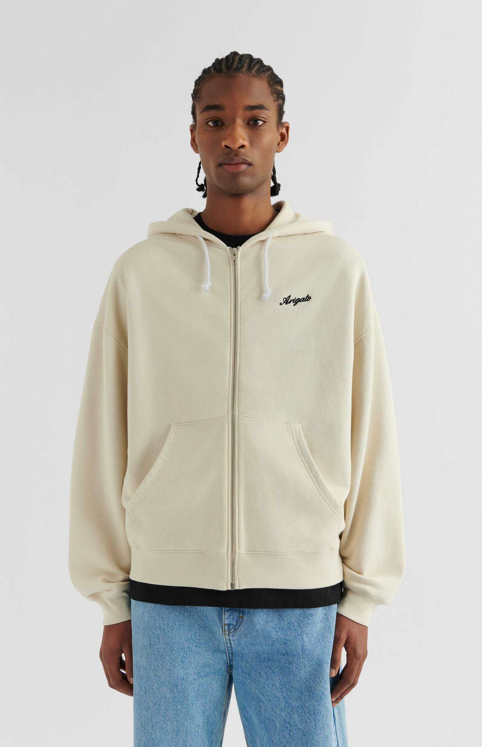 A young man with braided hair stands frontally, wearing an off-white zippered hoodie with 'Arigato' embroidered on the chest and light blue jeans. He is looking directly at the camera against a plain white background.