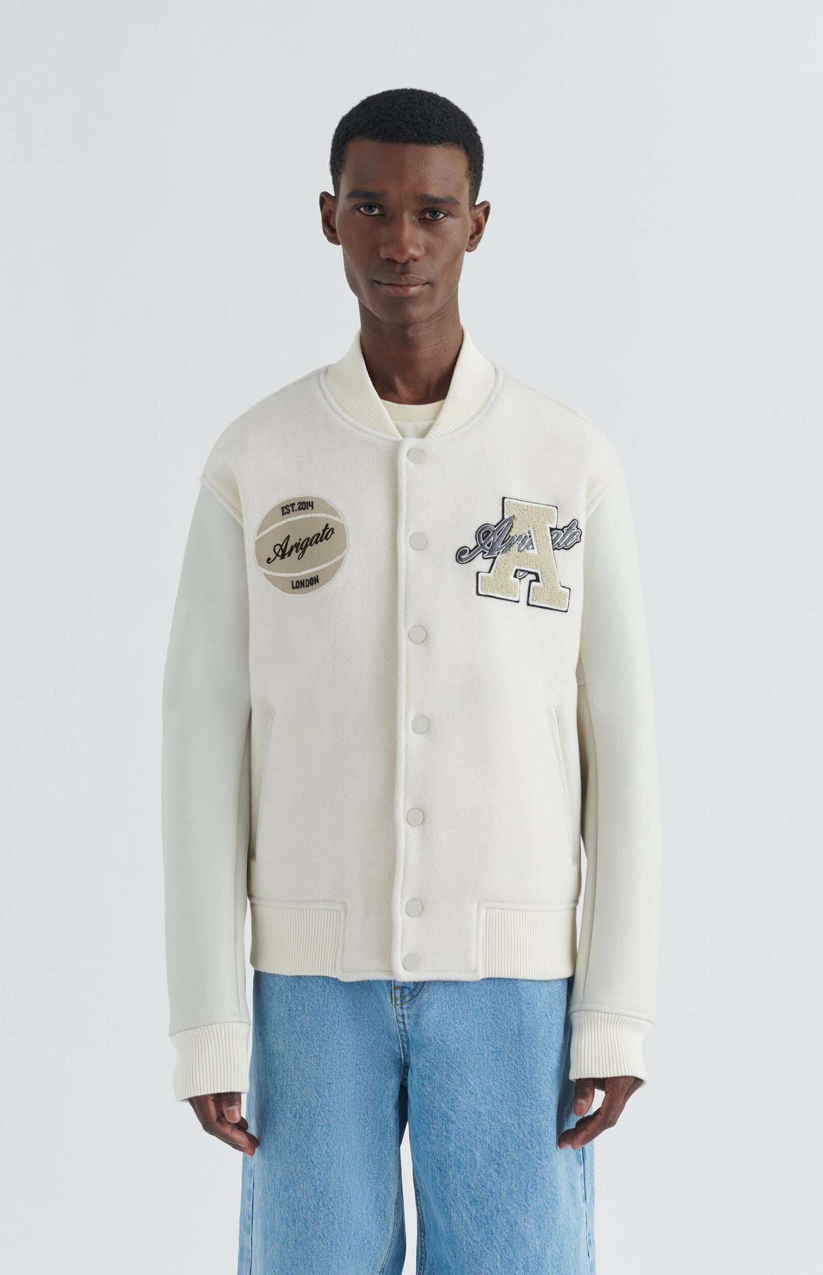 A man stands facing forward, wearing an off-white varsity jacket with a large 'A' patch and an 'Arigato London Est. 2014' circular patch, paired with light blue wide-leg jeans, against a plain white background.