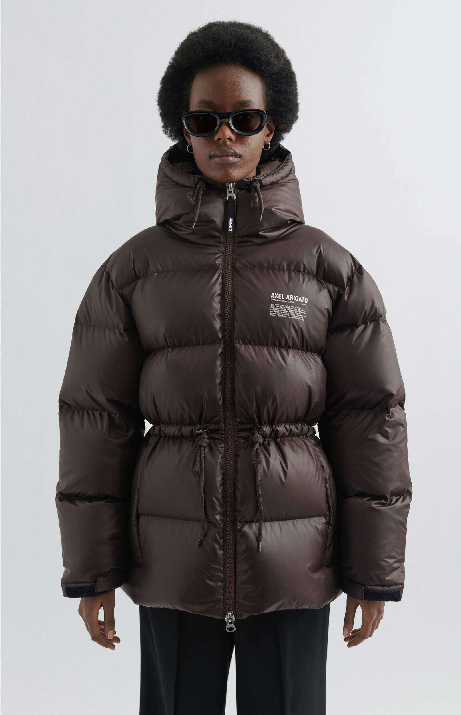 A person with dark curly hair and black sunglasses models a glossy dark brown puffer jacket with a hood, a zippered front, and a drawstring cinch at the waist. The brand 'AXEL ARIGATO' is visible in white text on the chest. The background is a plain light gray.