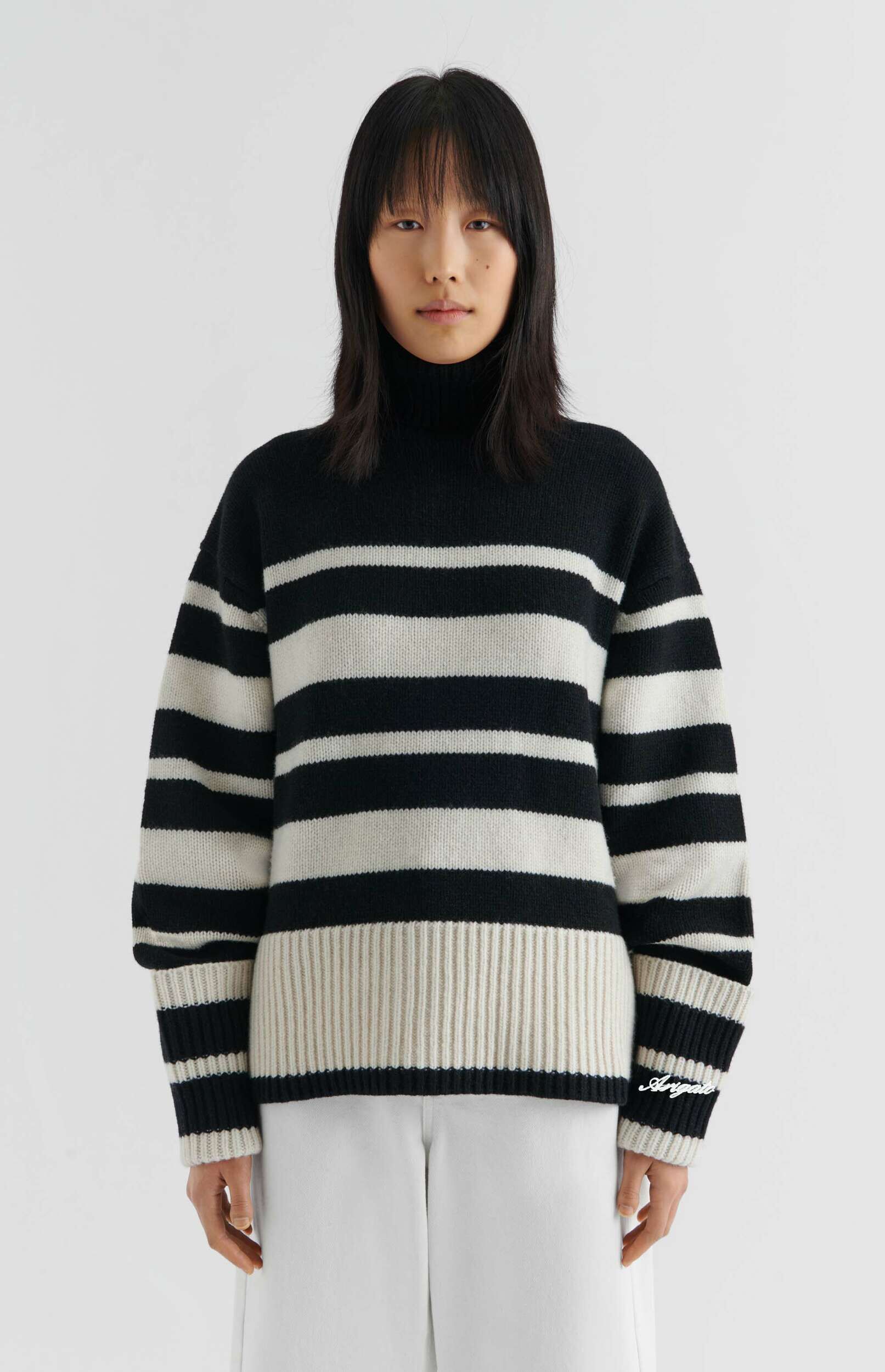 A person with dark hair and bangs stands against a white background, wearing a black and off-white striped turtleneck sweater and white wide-leg pants. The sweater features bold horizontal stripes, a high ribbed collar, and a wide ribbed off-white hem. The word 'Arigato' is embroidered in white script on the right sleeve cuff.
